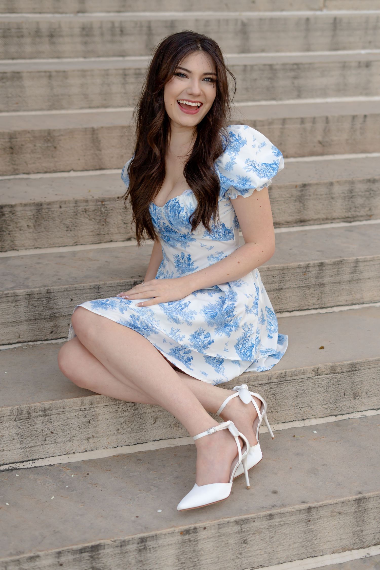 A woman in a blue and white floral dress with puff sleeves sits on concrete steps wearing white heels.