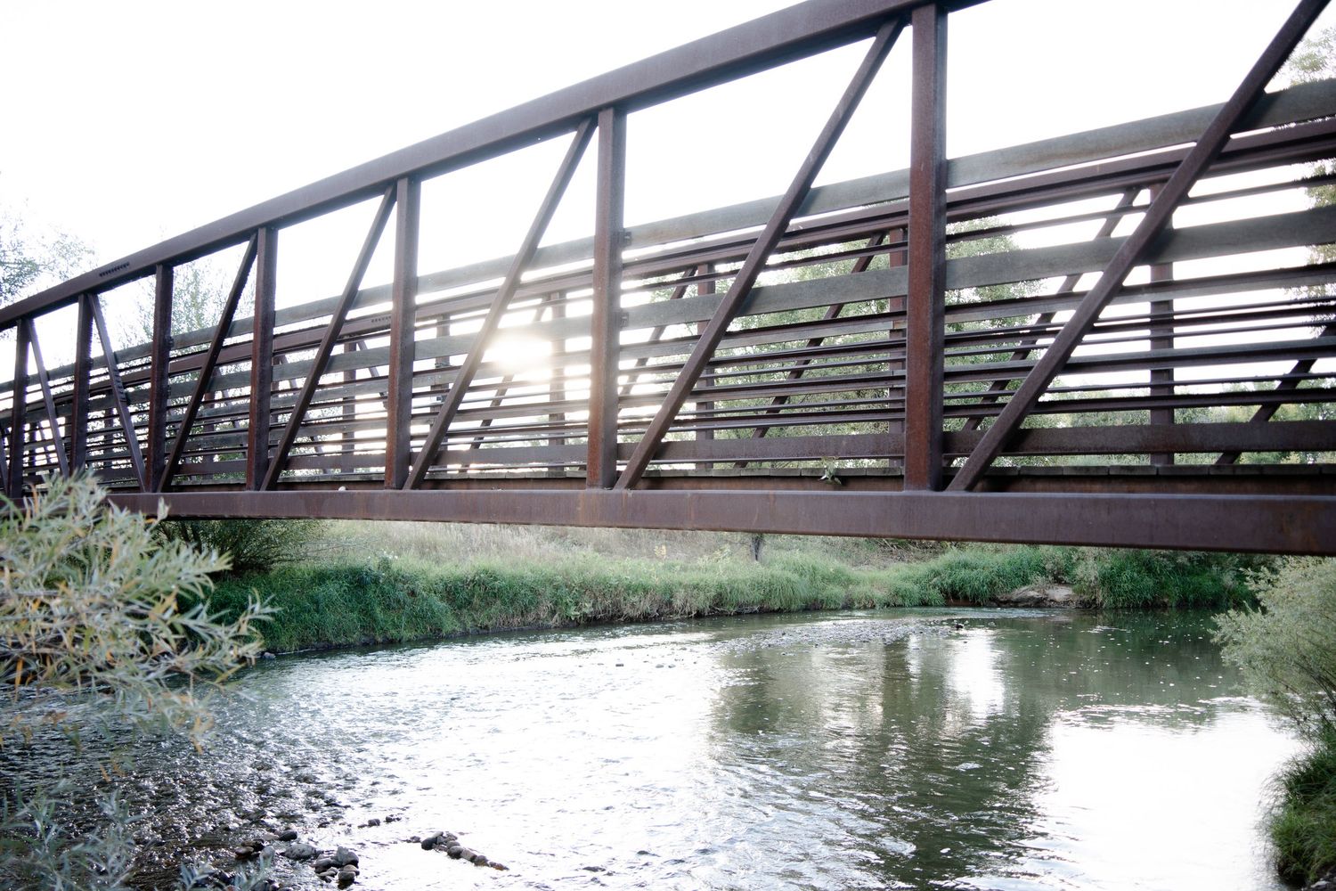 A rustic wooden footbridge spans across a tranquil river surrounded by lush green foliage and trees.