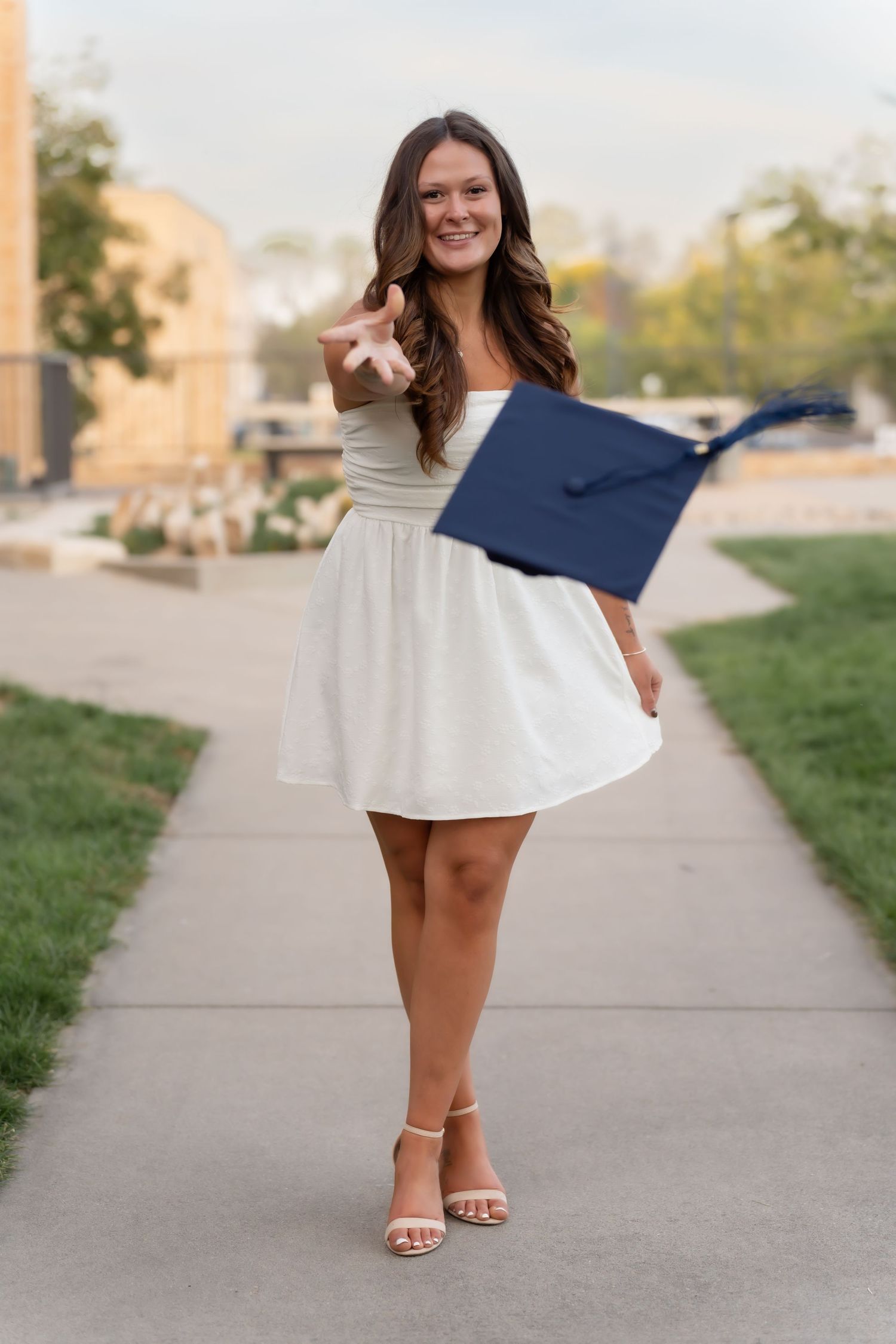 Graduate in white dress poses with diploma on campus sidewalk during sunset.