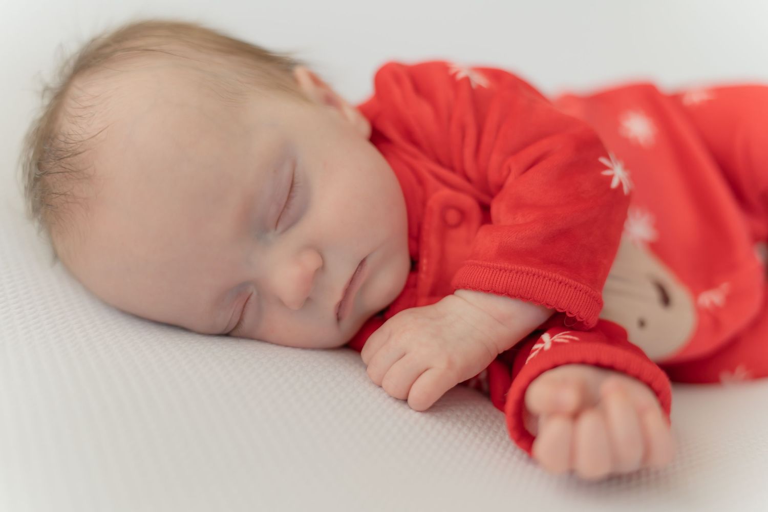 A sleeping baby in a red outfit rests peacefully on a white blanket.