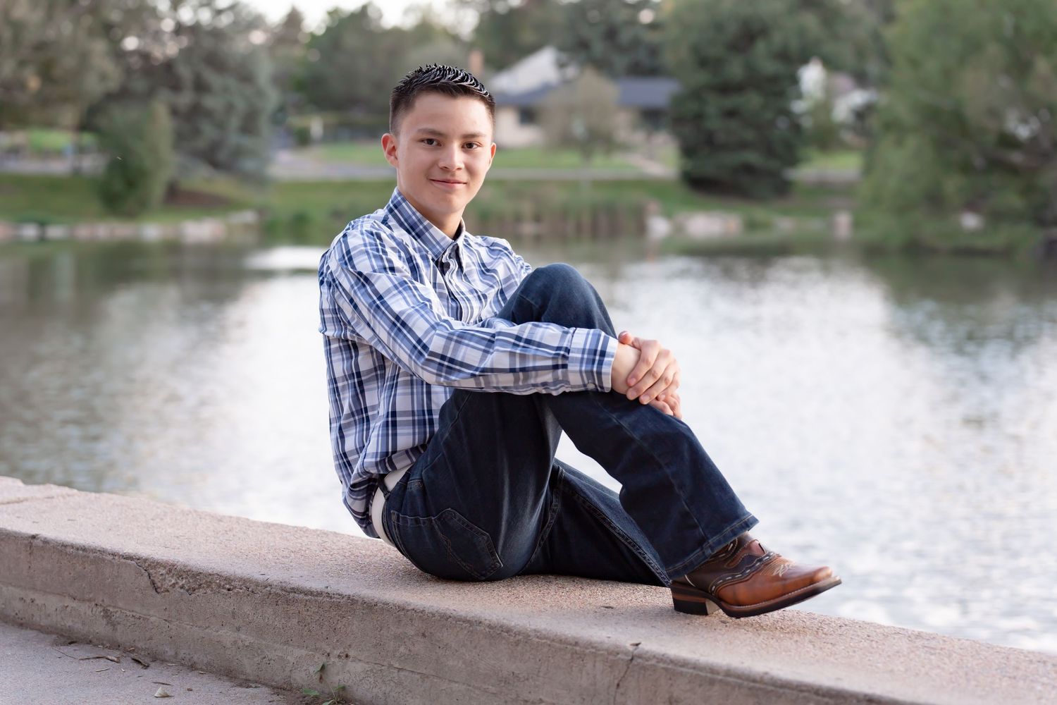 Student in plaid shirt and jeans sits casually on wall by lake.