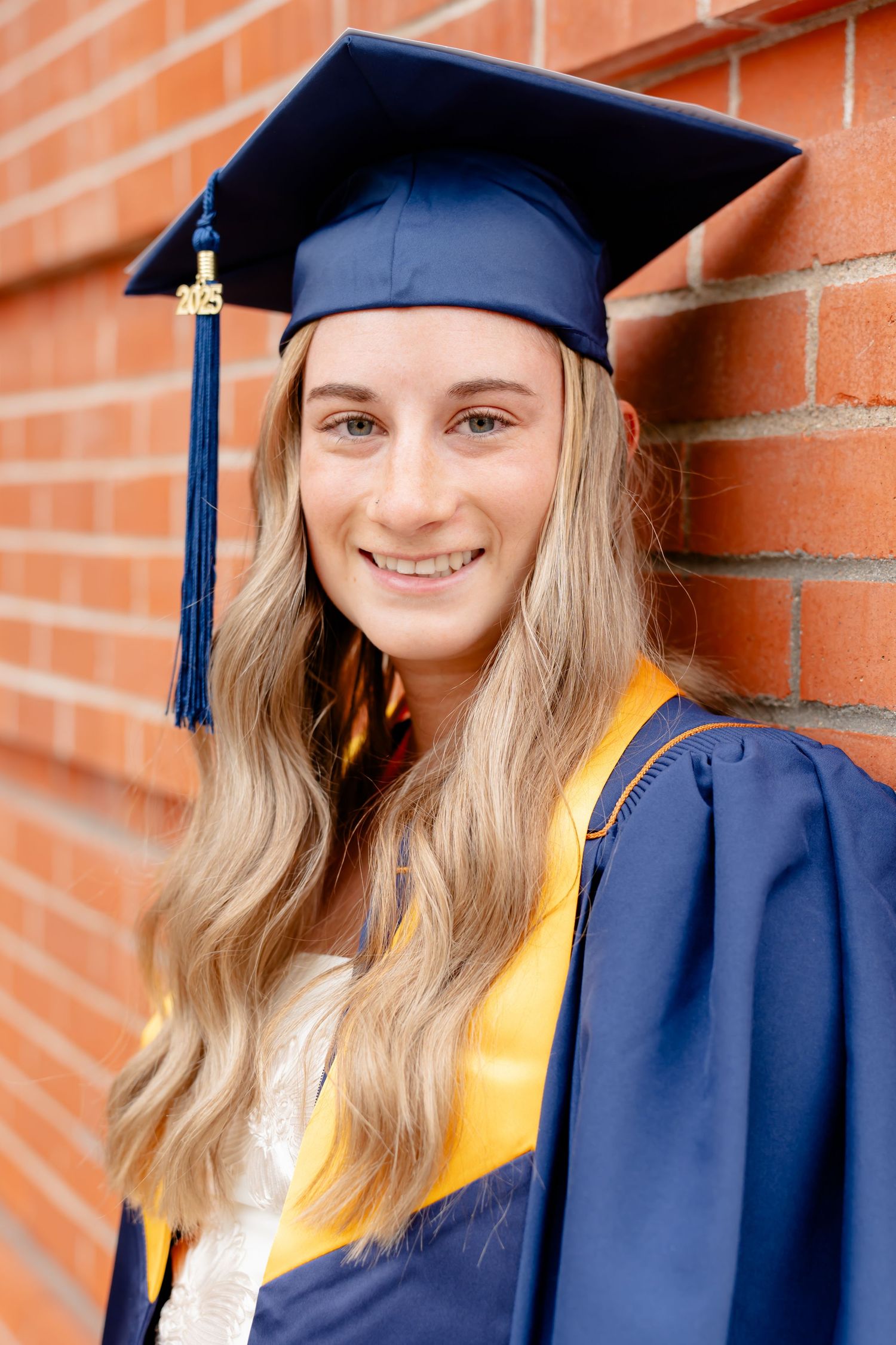 Graduate in navy cap and gown with gold stole poses against brick wall background.