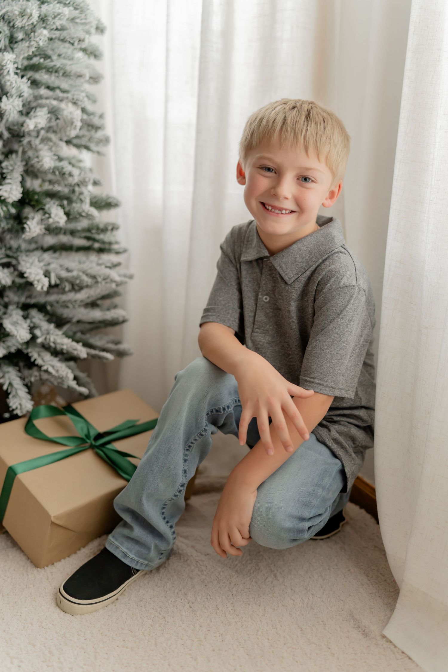 Child in gray polo shirt sits by Christmas tree with wrapped present on carpeted floor.