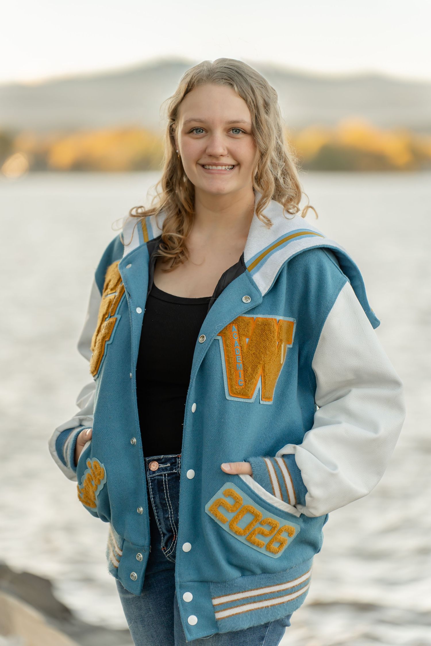 Person wearing a blue and gold varsity letterman jacket poses by water during a photo session.