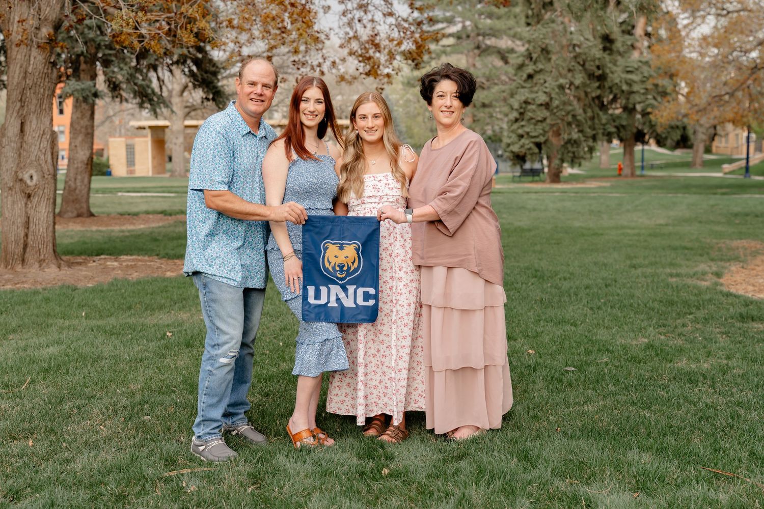 Family group poses outdoors holding UNC banner on grassy campus lawn.