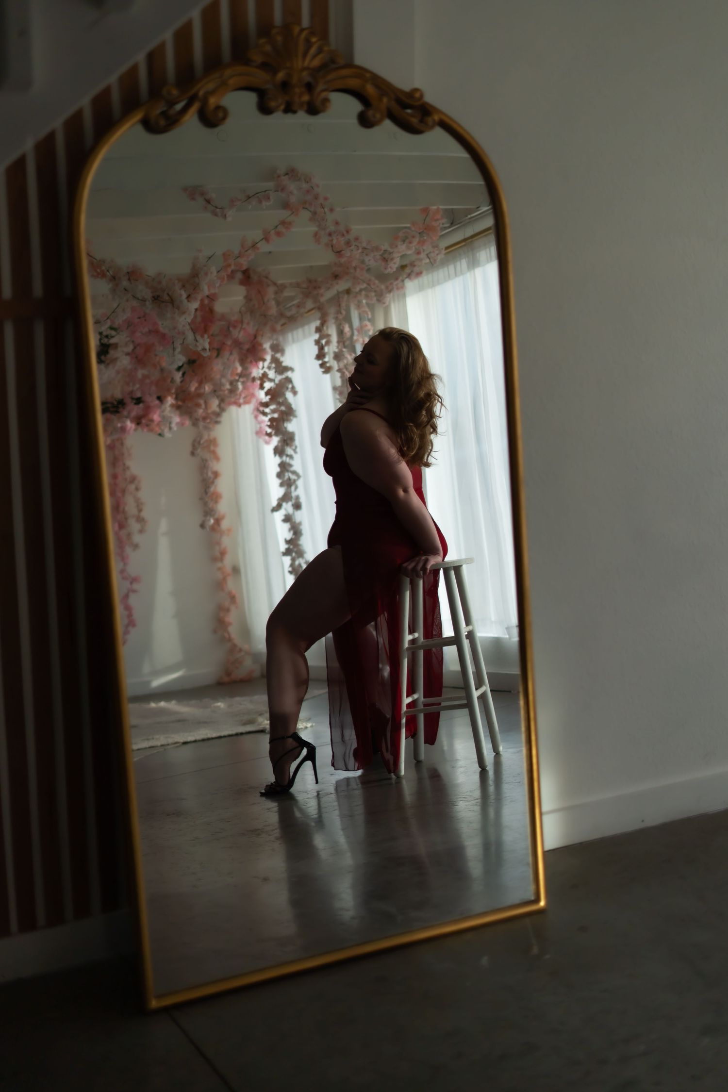 A darkly lit room with a gold-framed mirror reflects a silhouette sitting on a chair against floral wall decorations.