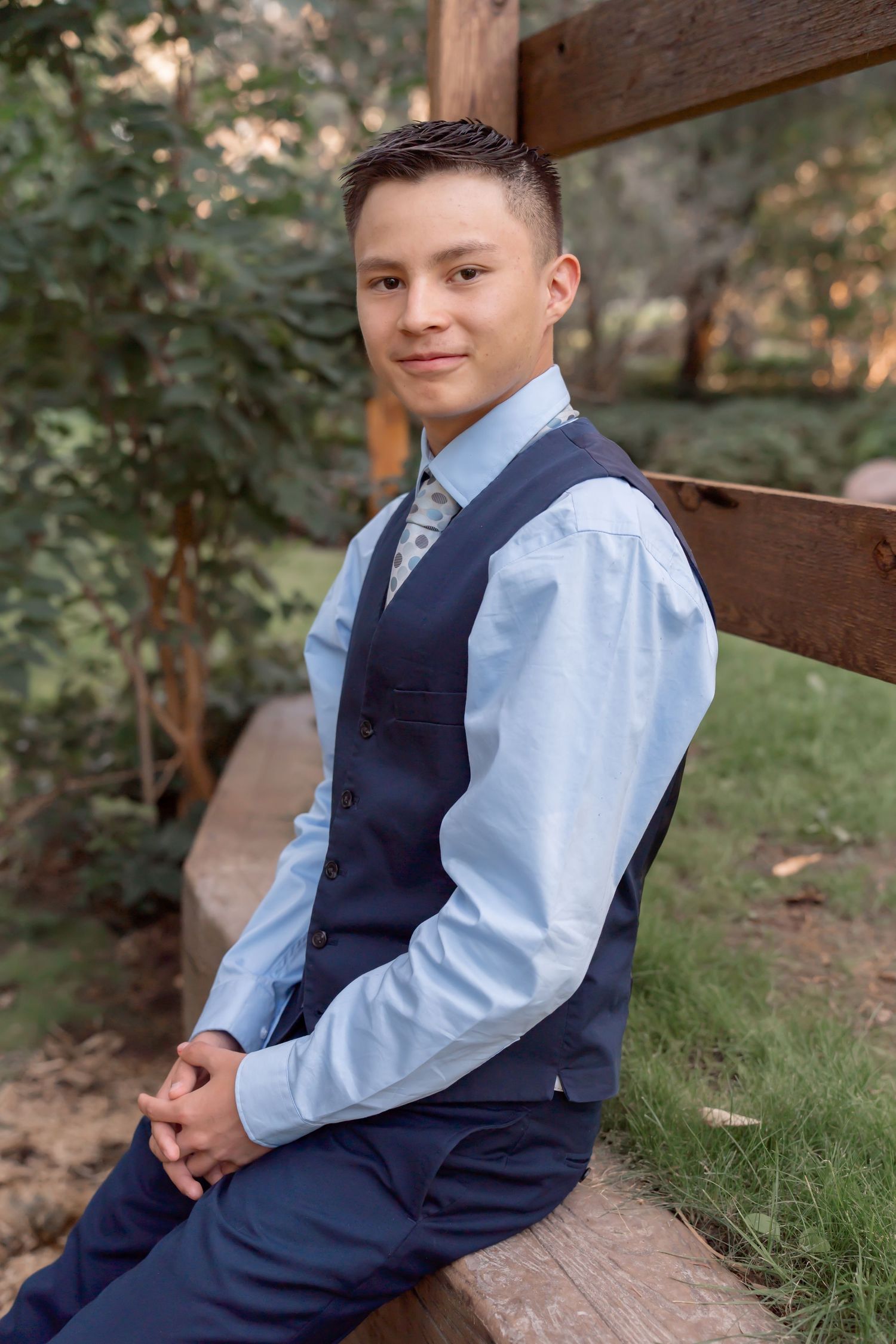 A person in business attire sits on a wooden fence wearing a navy vest and light blue dress shirt.