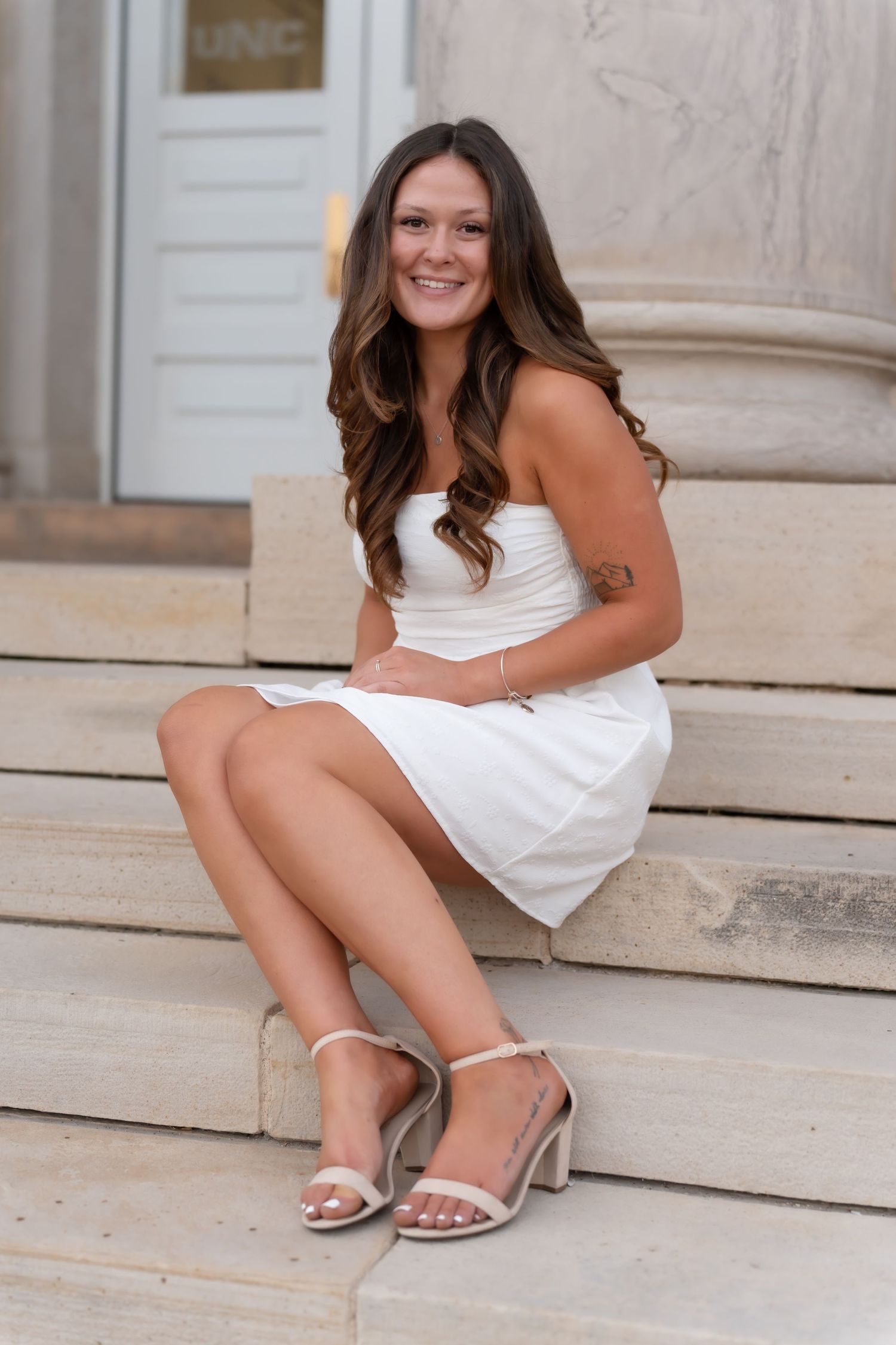 A person in a strapless white dress sits on stone steps, wearing silver sandals and smiling in an outdoor setting.
