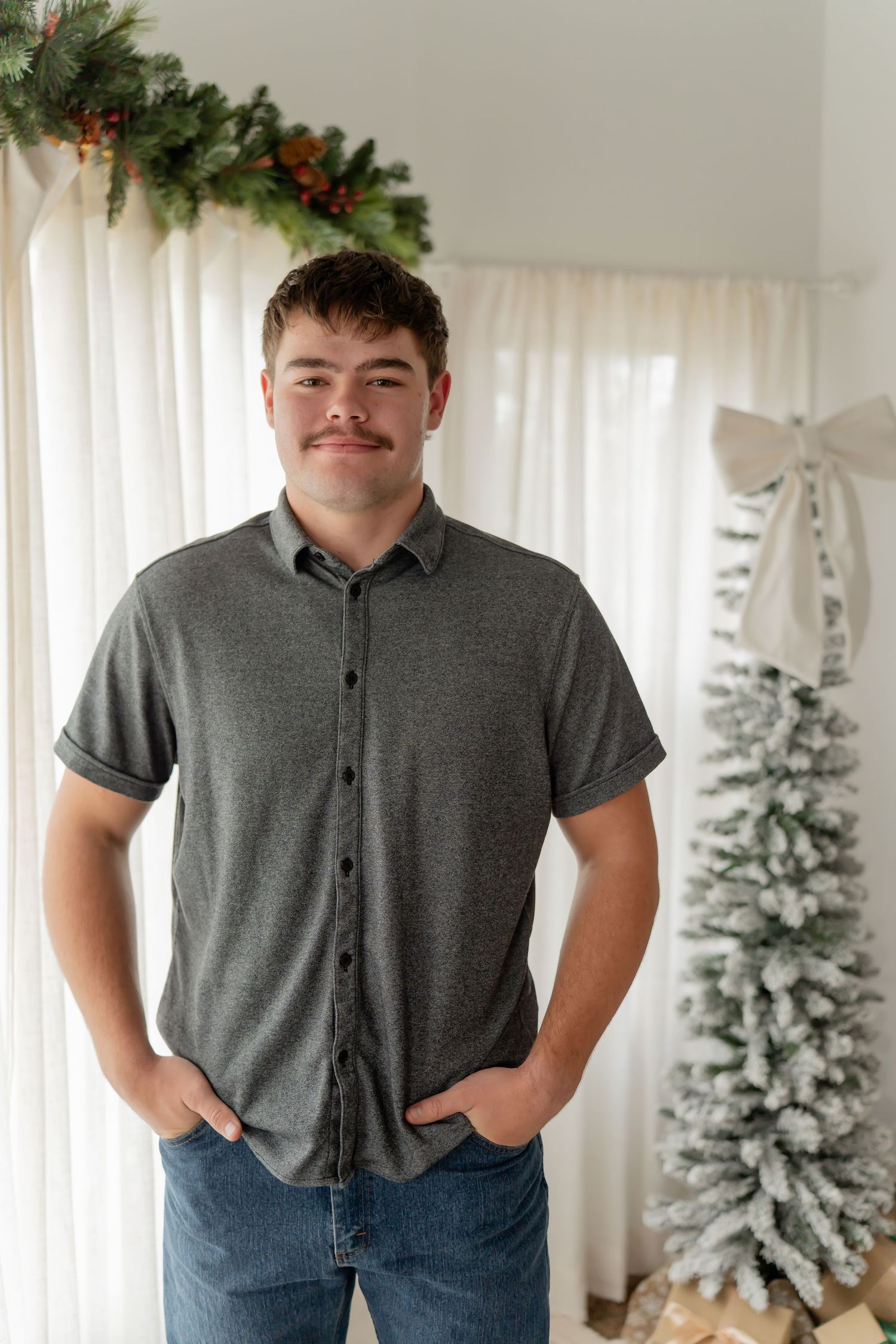 Someone poses in a gray polo shirt and jeans in front of a white Christmas tree with festive decorations.