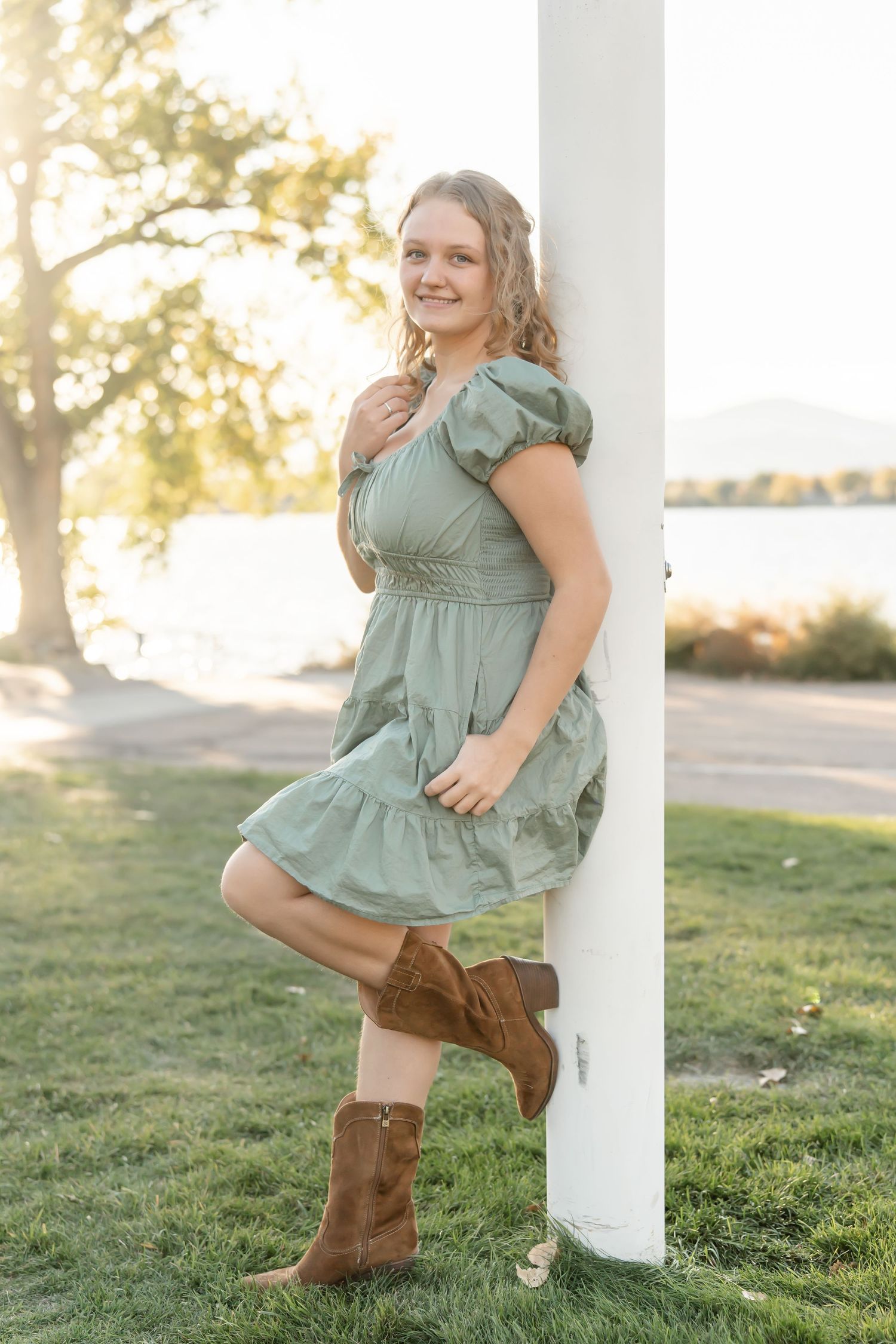 A person in a sage green dress and brown boots dances playfully by a white pillar in golden sunlight.