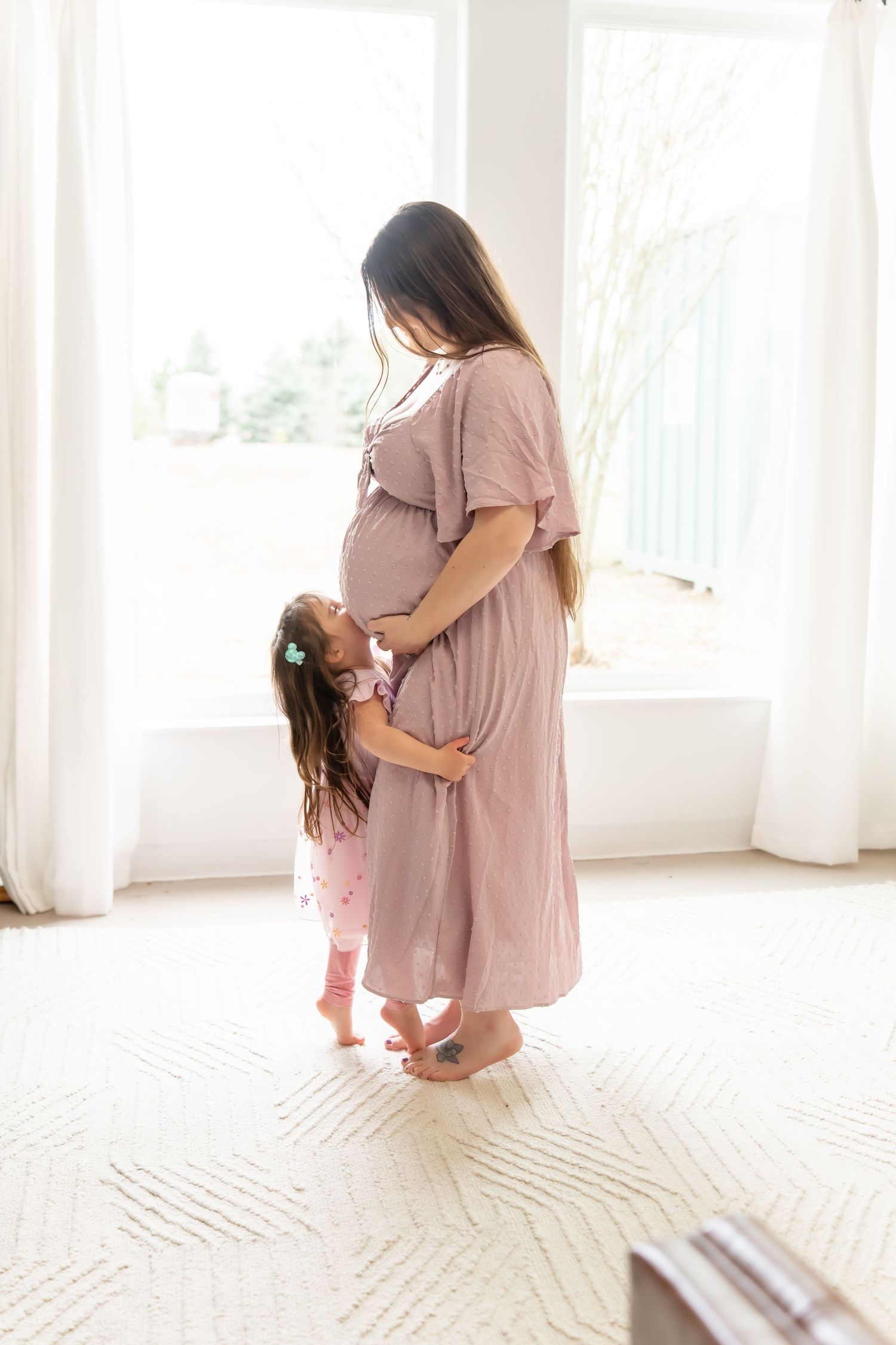 Mother and daughter share tender moments in a bright sunlit room with white curtains and carpet.