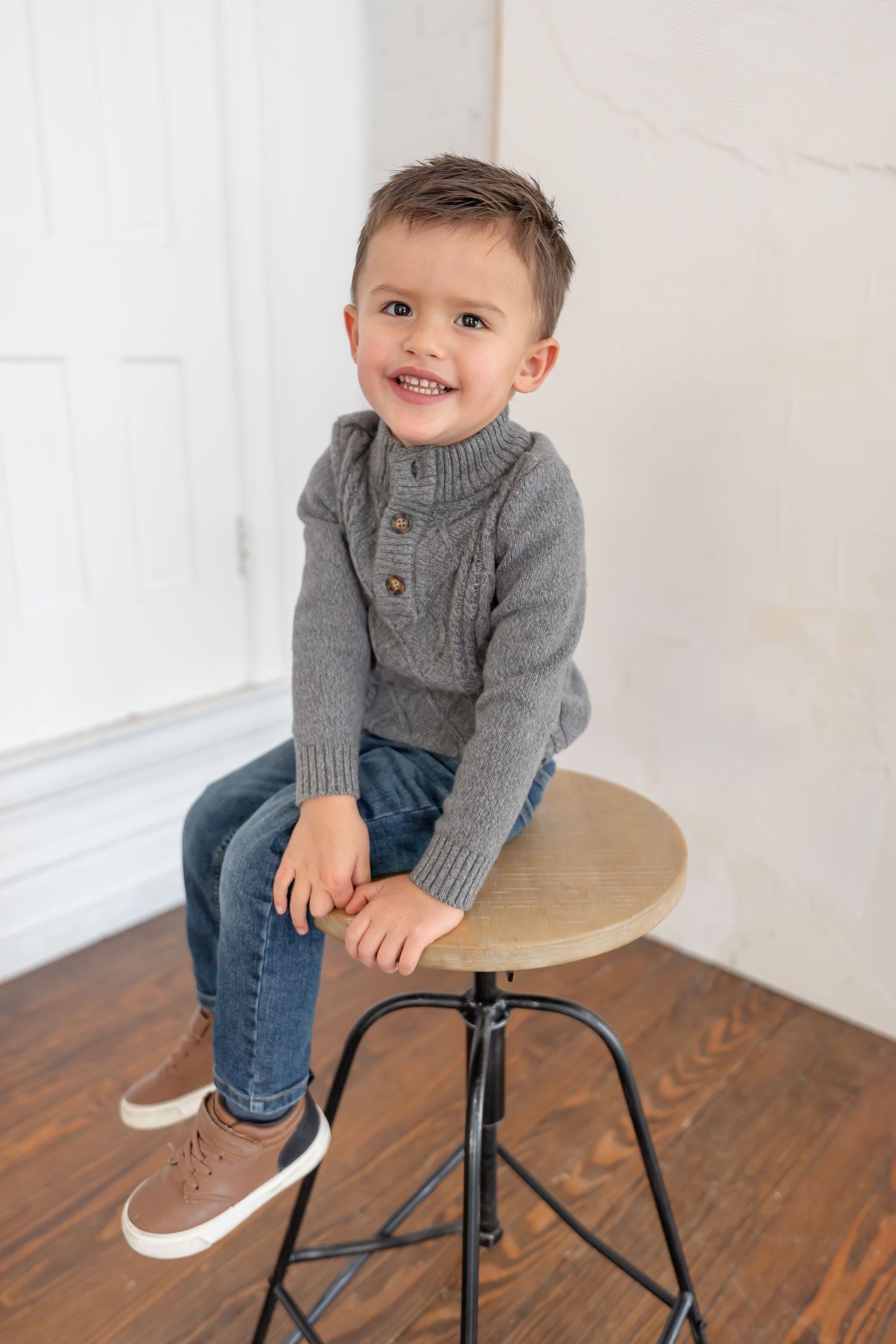 Child in grey sweater and jeans sits playfully on wooden stool against white wall in natural light setting.