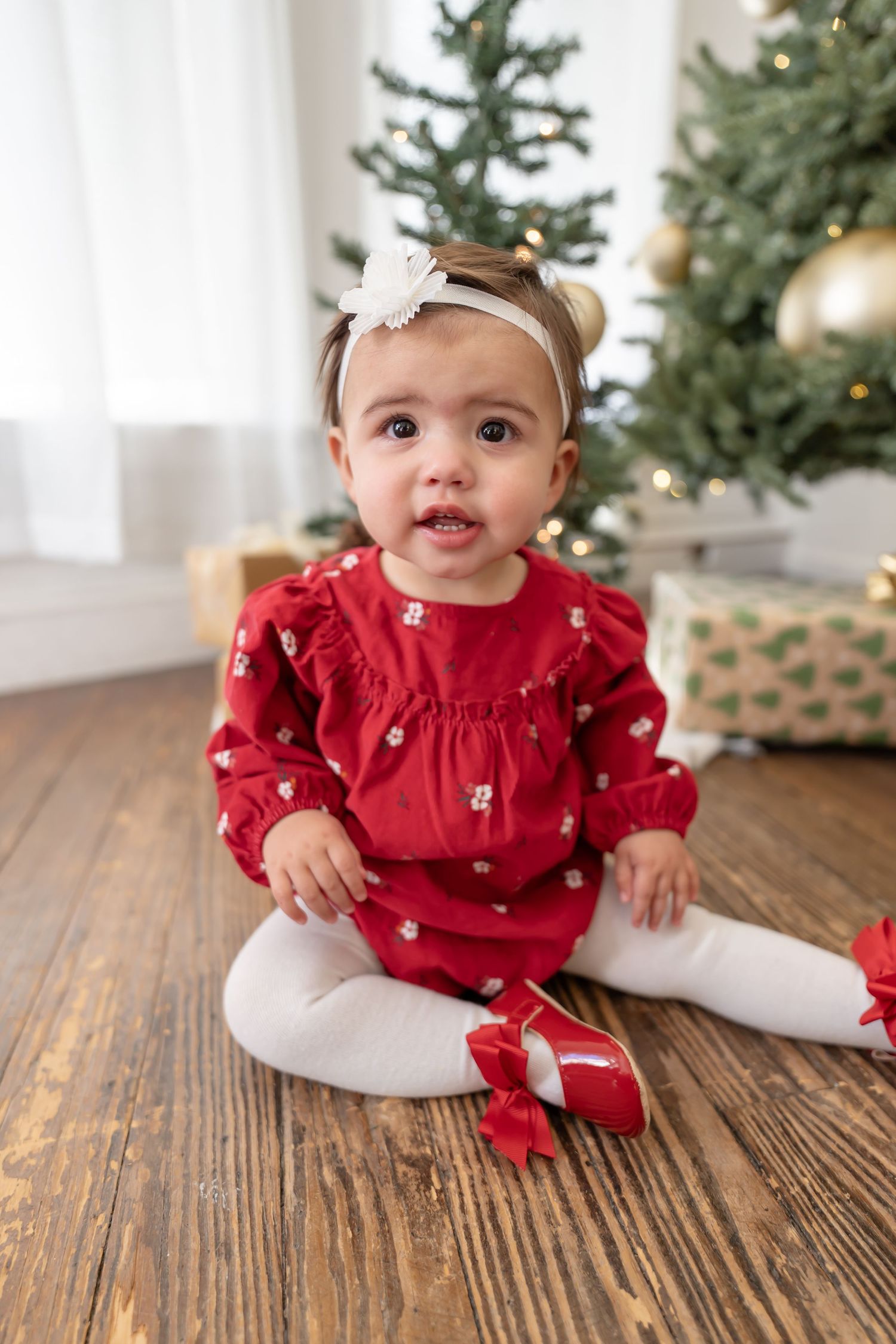 Baby in red holiday dress sits on wooden floor near Christmas tree with festive decorations.