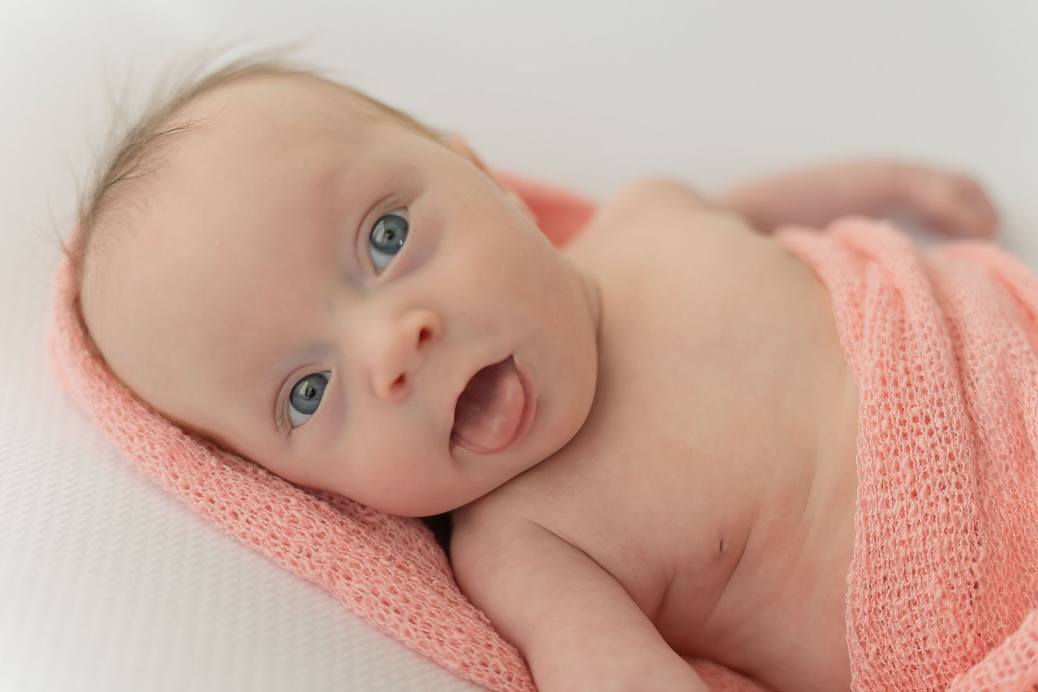 A baby wearing a soft pink knit wrap lies on white bedding while looking at the camera with bright blue eyes.