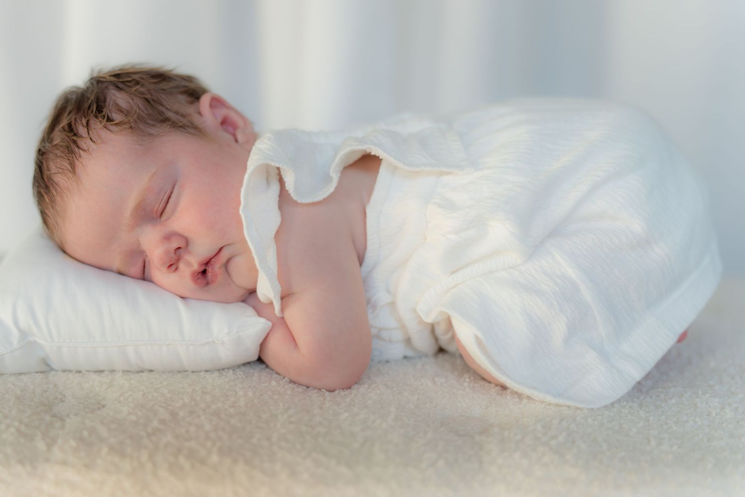 Sleeping baby in white dress on white pillow.