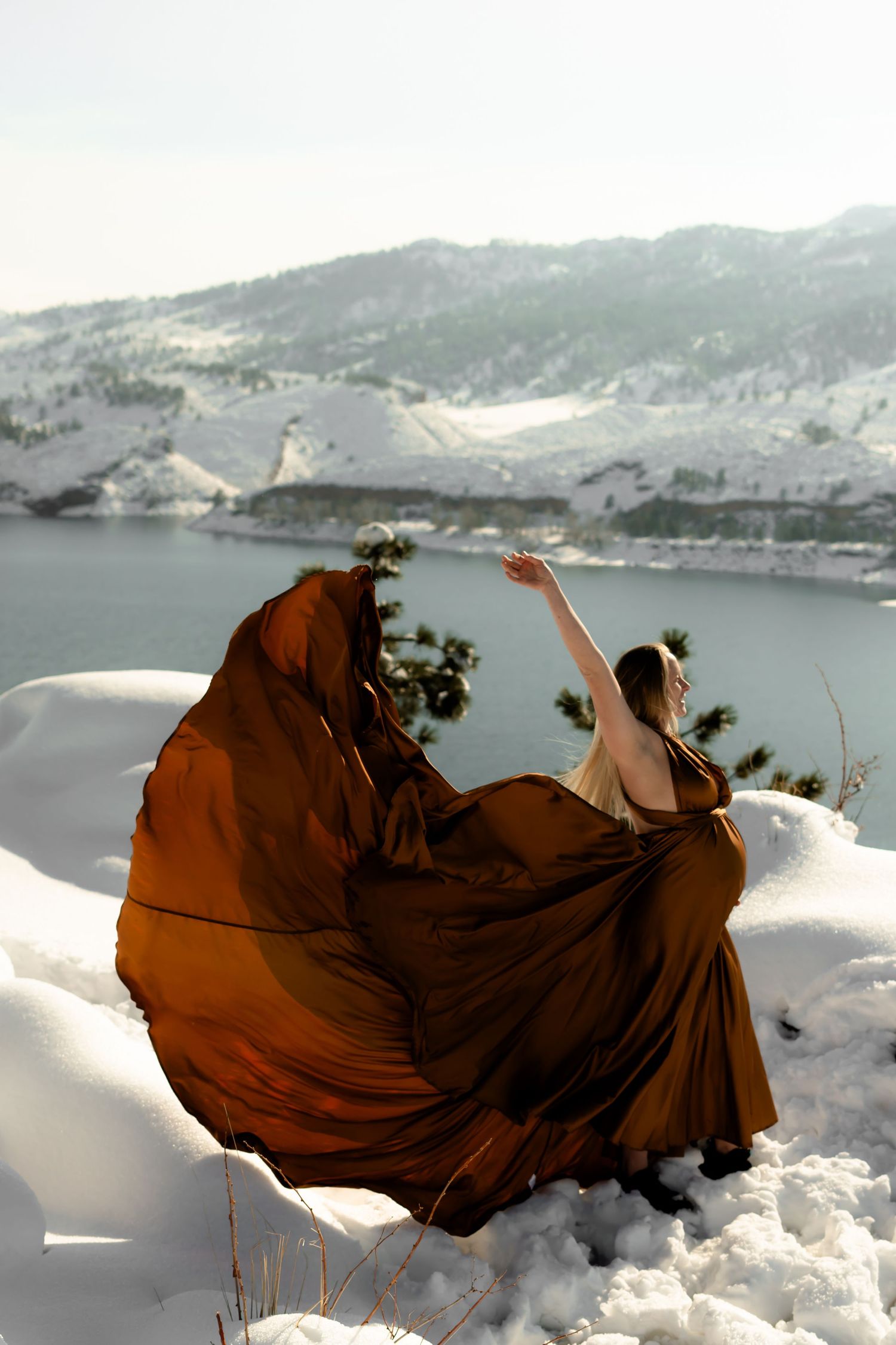 Brown dress billows dramatically in winter mountain landscape with snow and lake view.