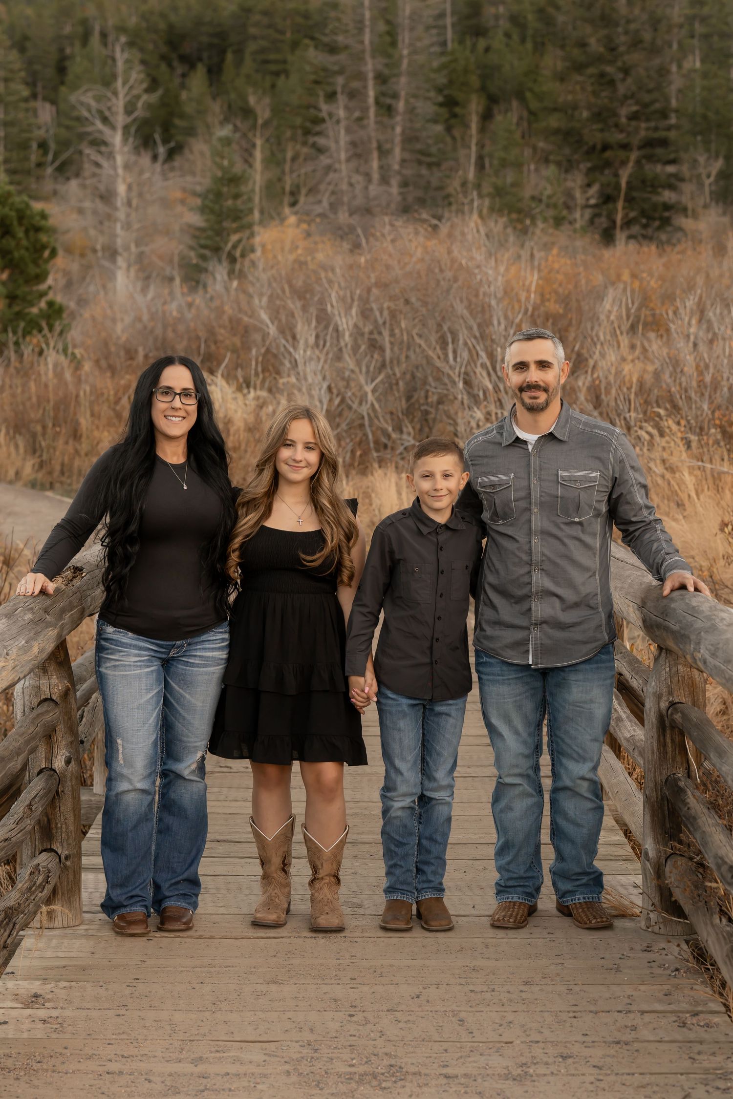 A family group stands together on a wooden bridge path in an autumn outdoor setting with bare trees in the background.
