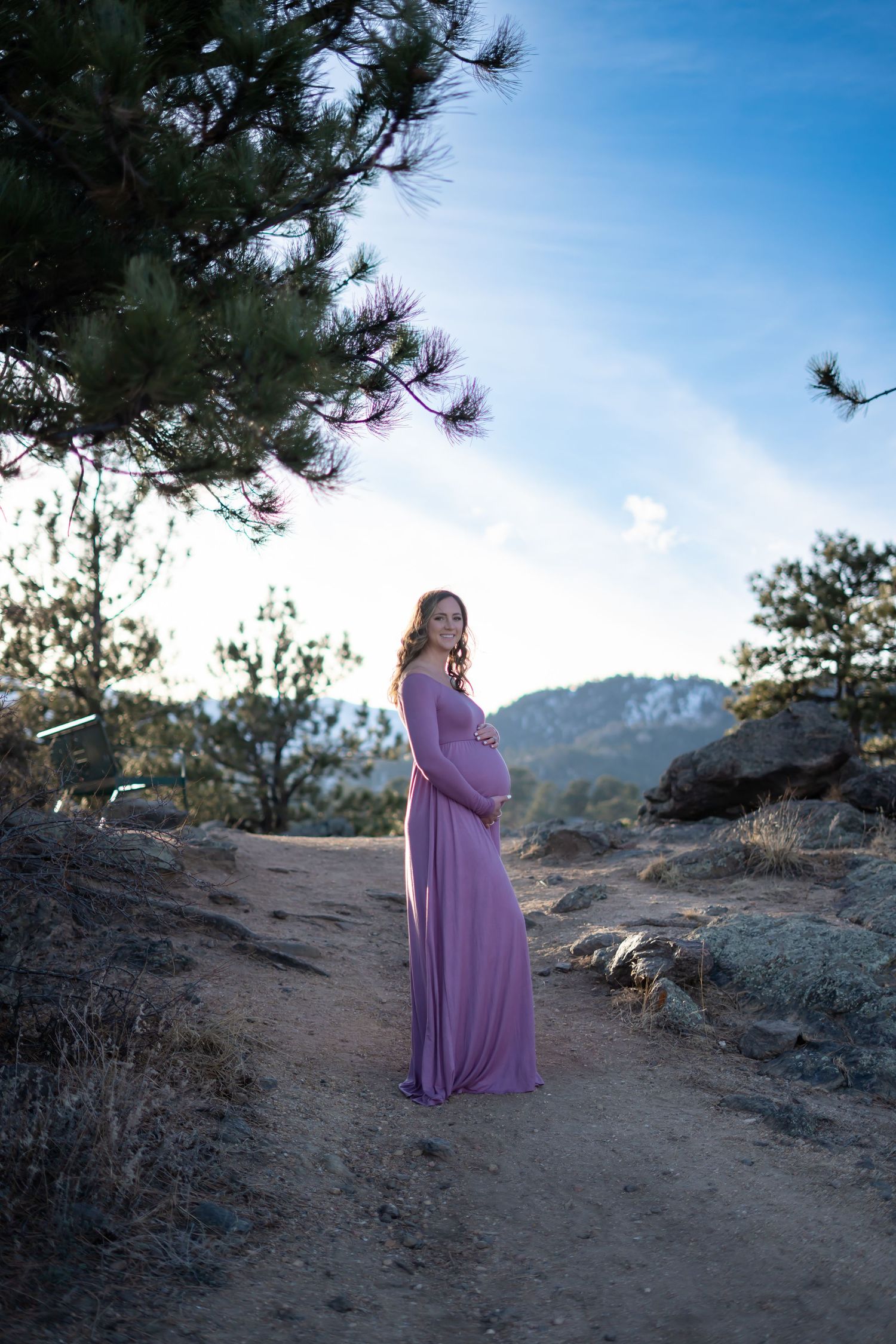 A silhouetted figure in a flowing purple dress stands on a mountain trail at sunset.