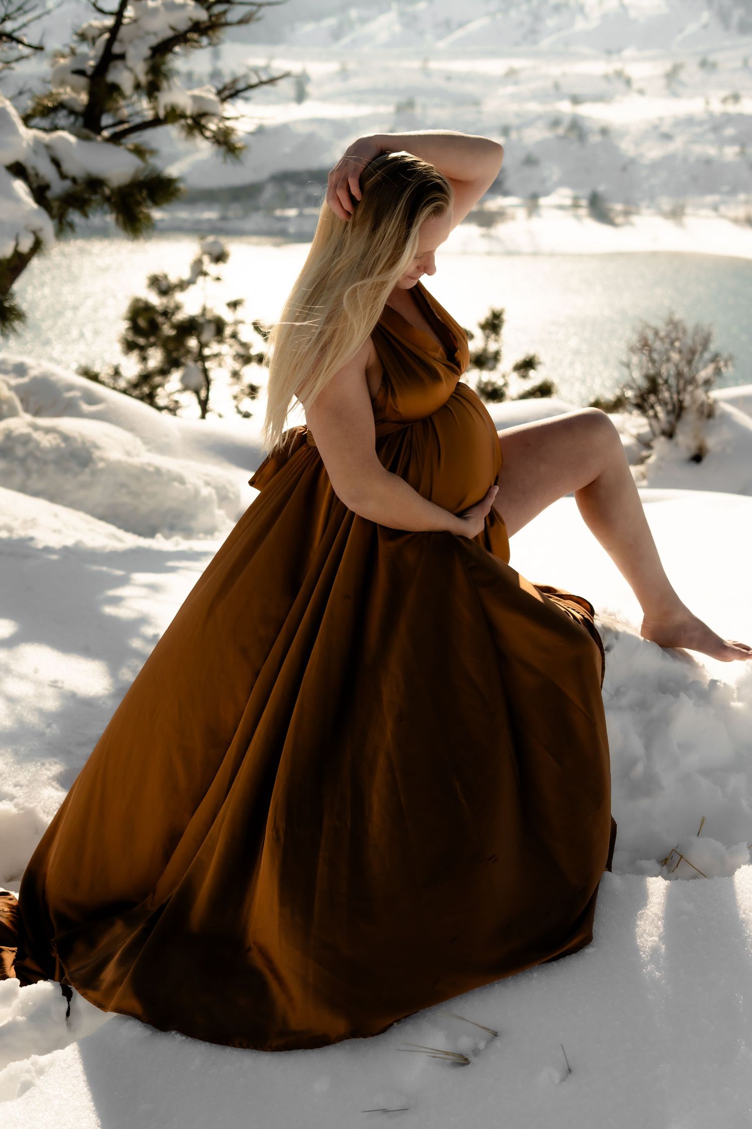 A flowing brown dress creates a dramatic silhouette against snowy mountain landscape during maternity photoshoot.