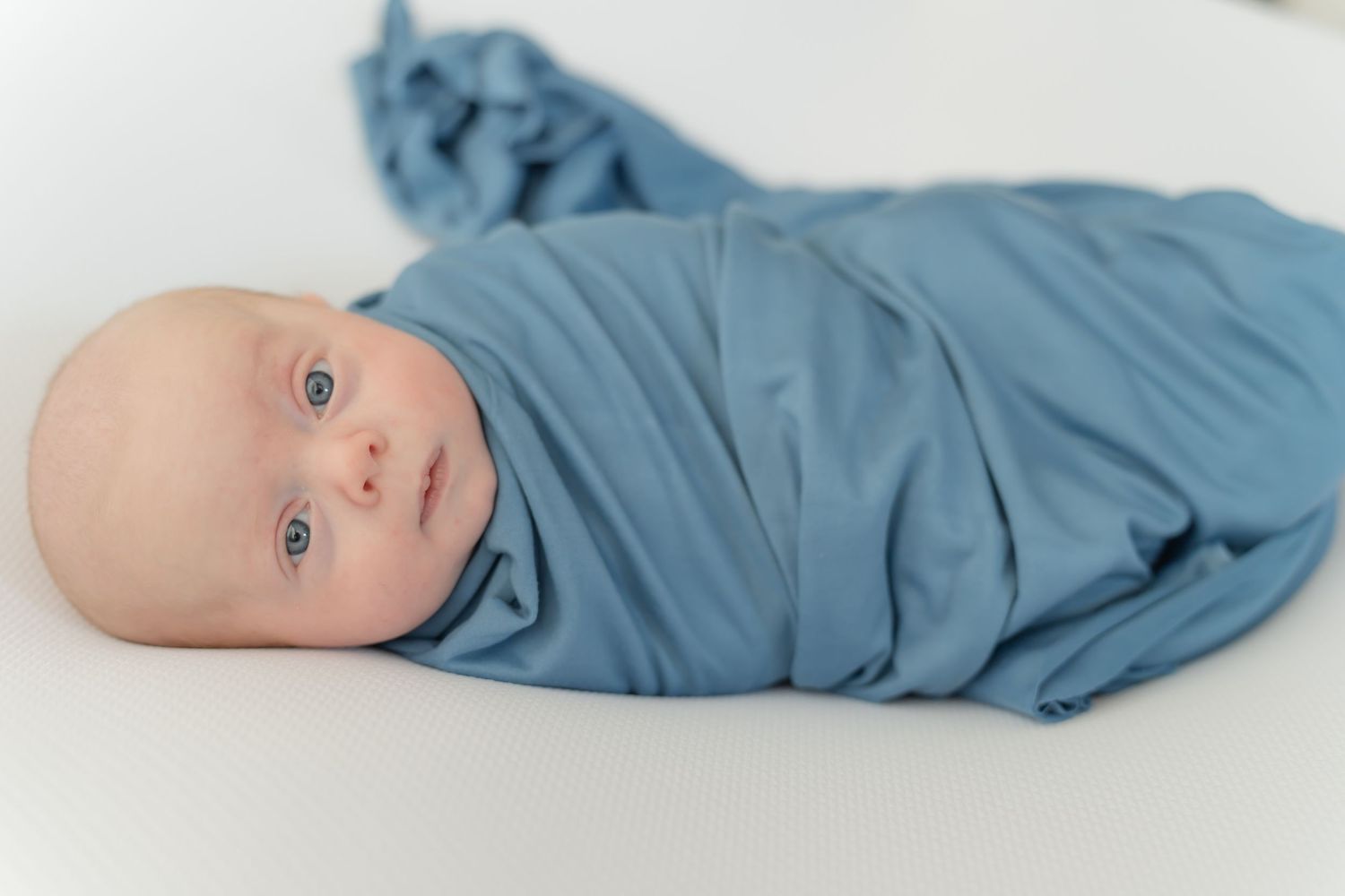 A baby wrapped in soft blue fabric lies on a white background in a creative photo series.