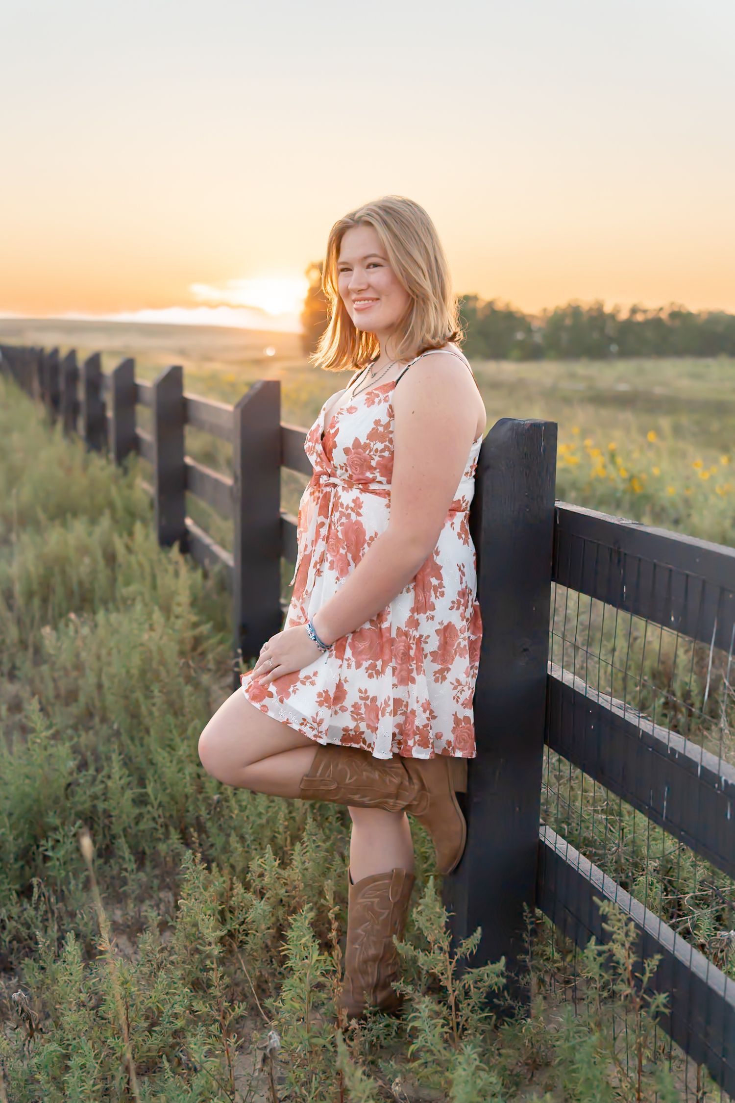 A person in a floral top and brown pants poses against a black fence at sunset in a rural field.