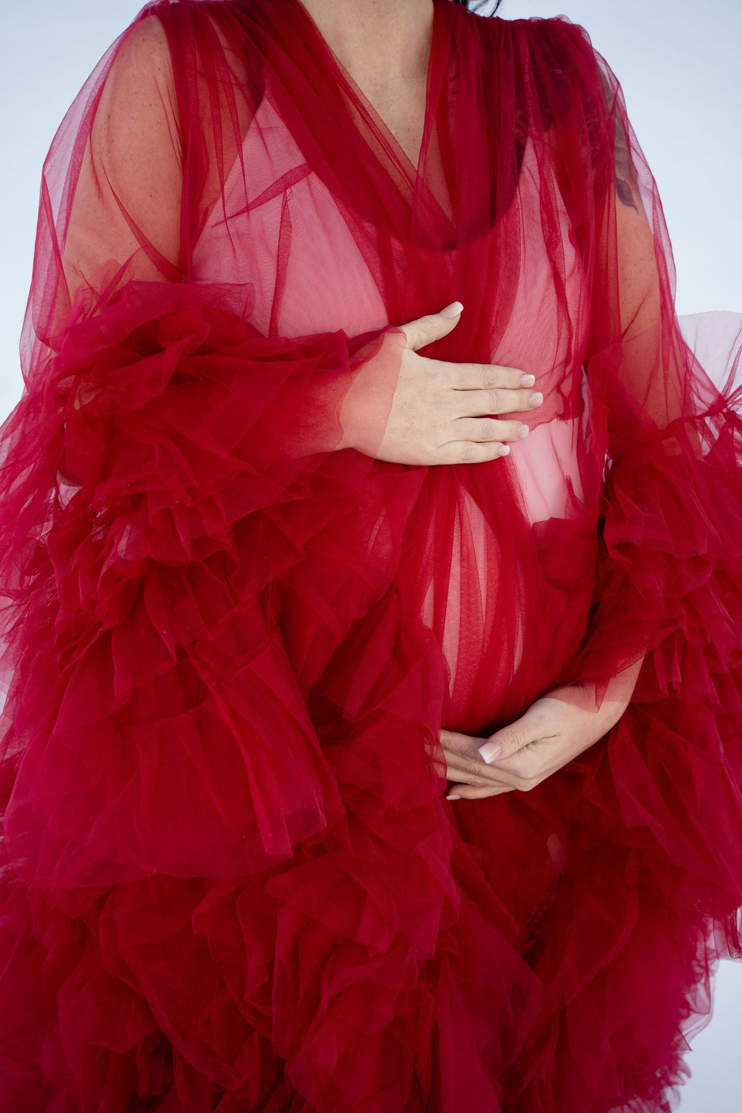 Close-up detail of red tulle maternity gown with hands placed on belly.