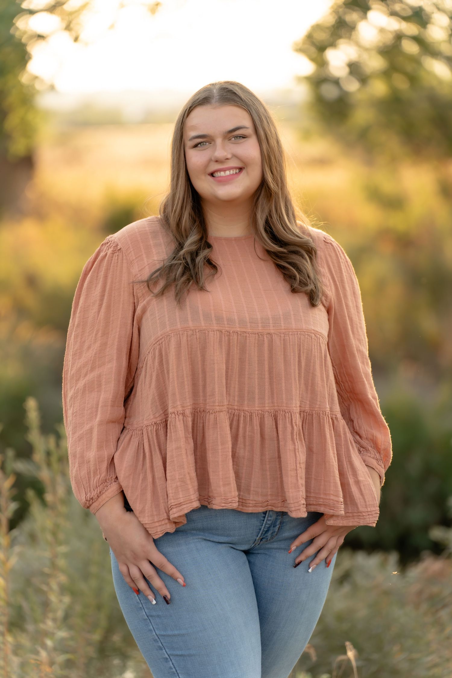 A portrait in a peach-colored ruffled top and jeans against a golden sunset background.