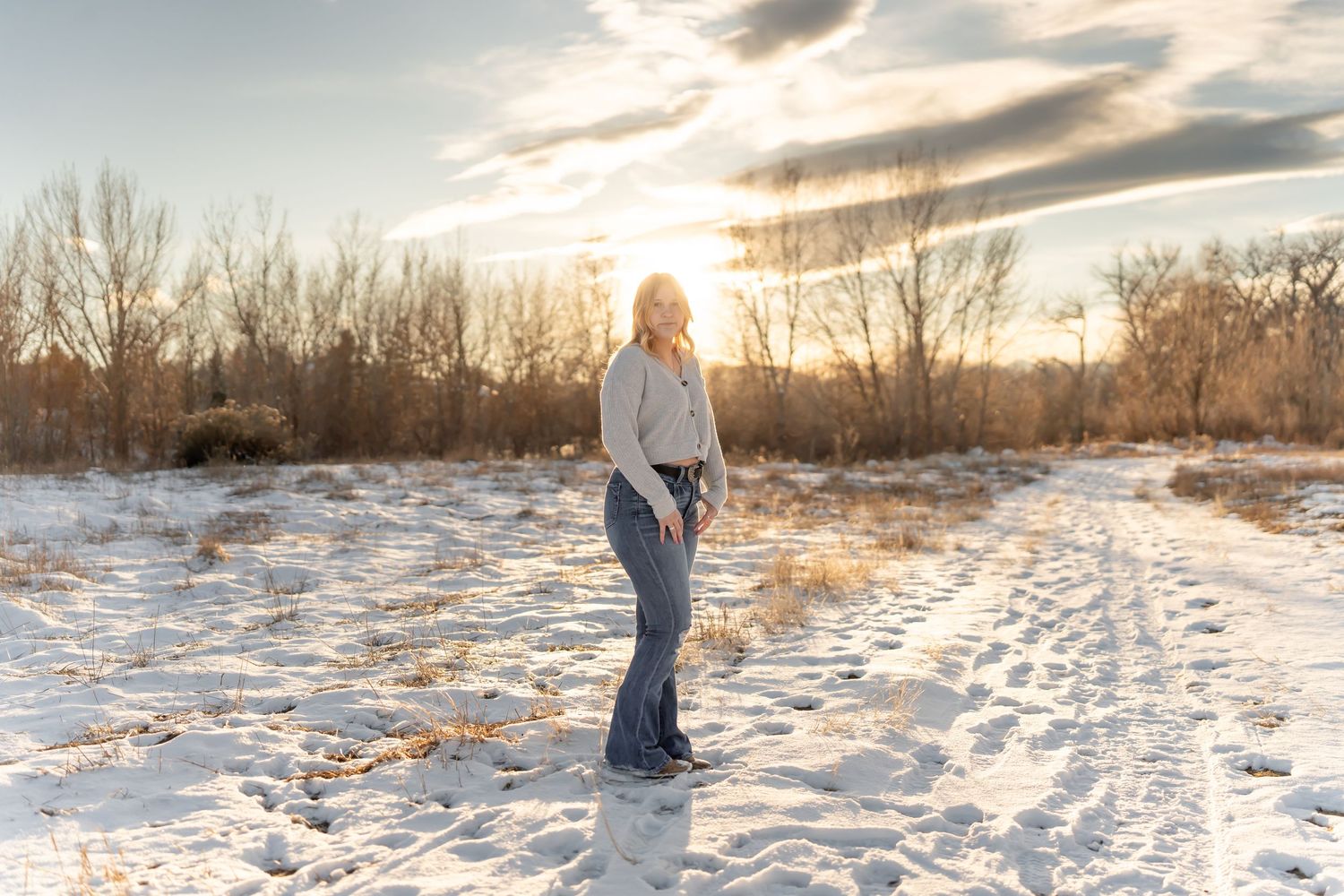 Person stands in snowy field at sunset wearing light colored sweater and jeans.