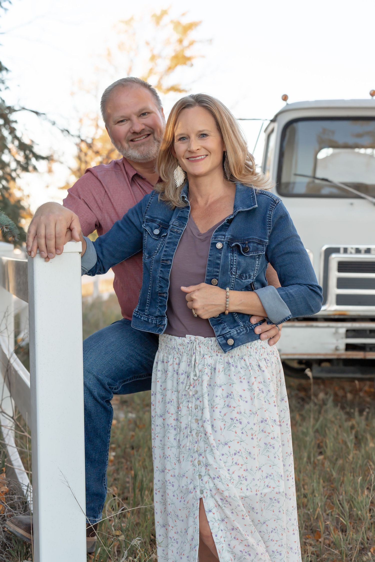 A couple posing together by a white fence with a vintage truck in the background.