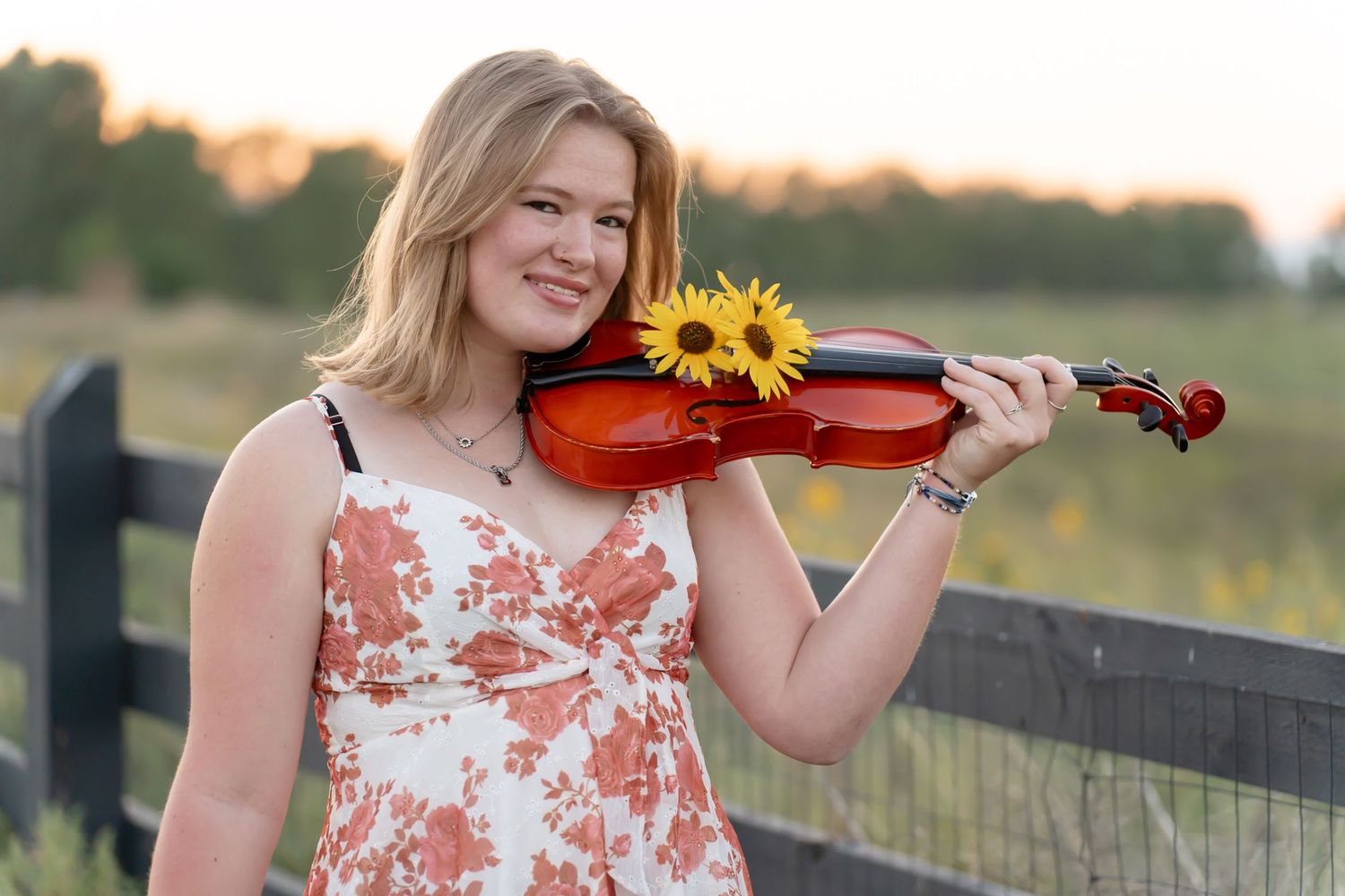 A person in a floral dress plays violin outdoors during golden hour in a rustic country setting.