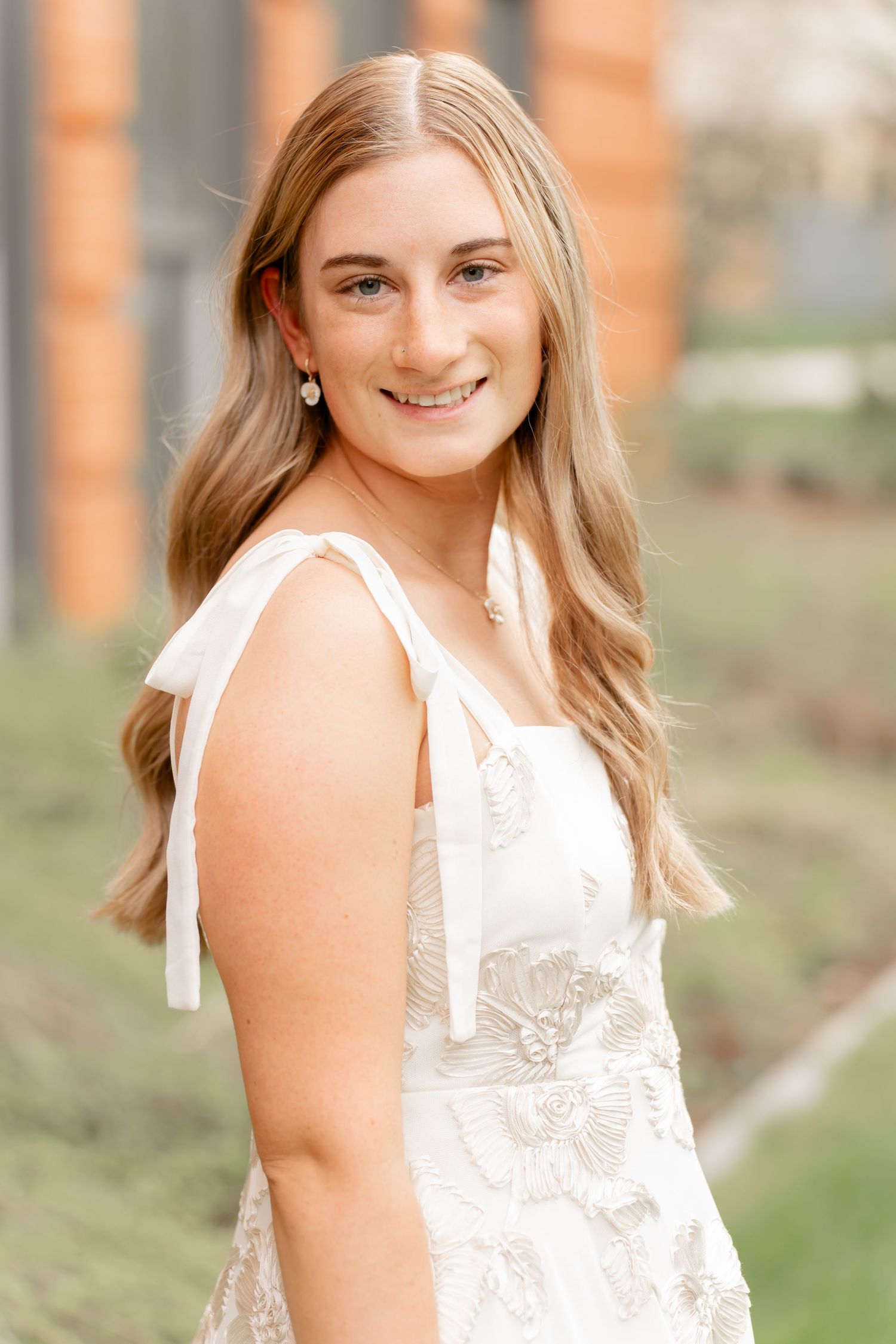 Portrait in white lace dress with shoulder ties against brick building background.