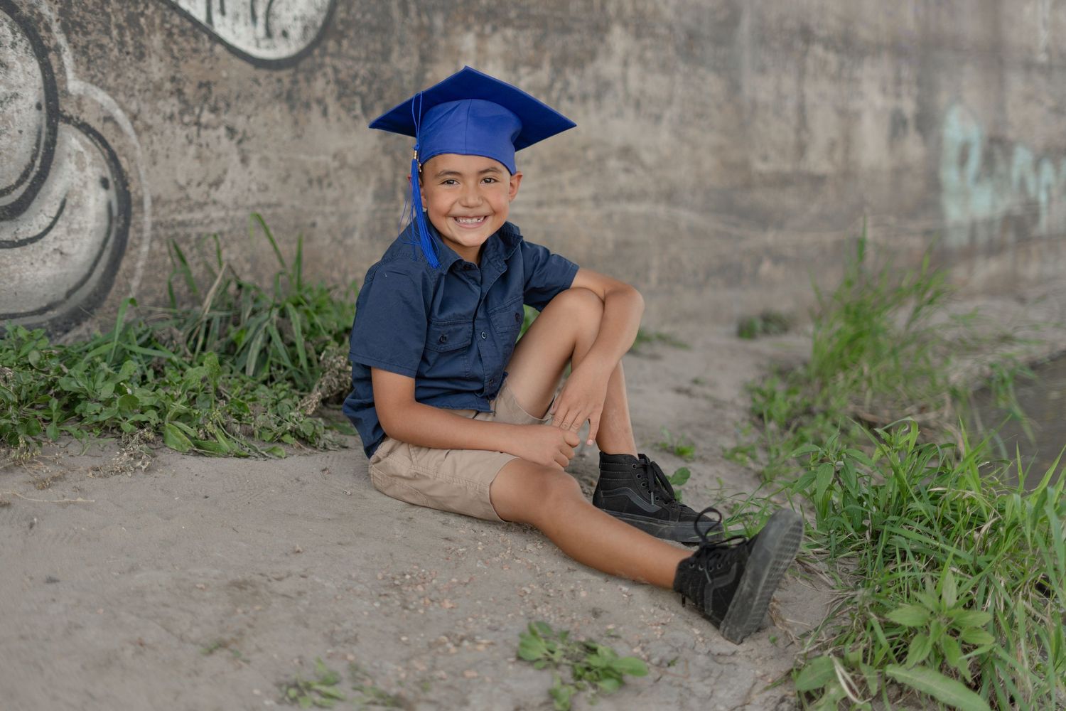 Graduate in blue cap and gown posing outdoors against stone wall backdrop.