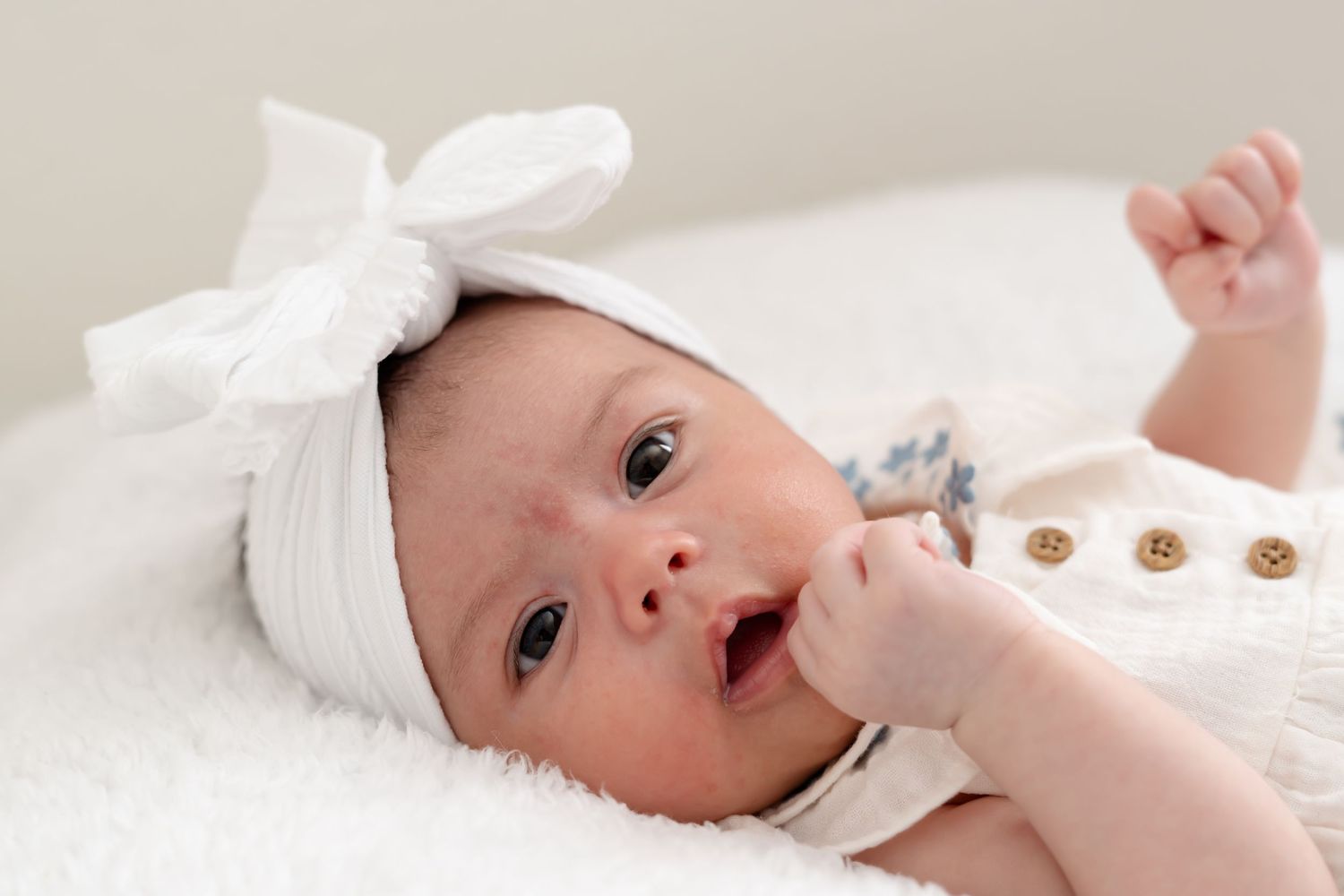 Baby wearing white bow headband looking up at camera with curious expression.