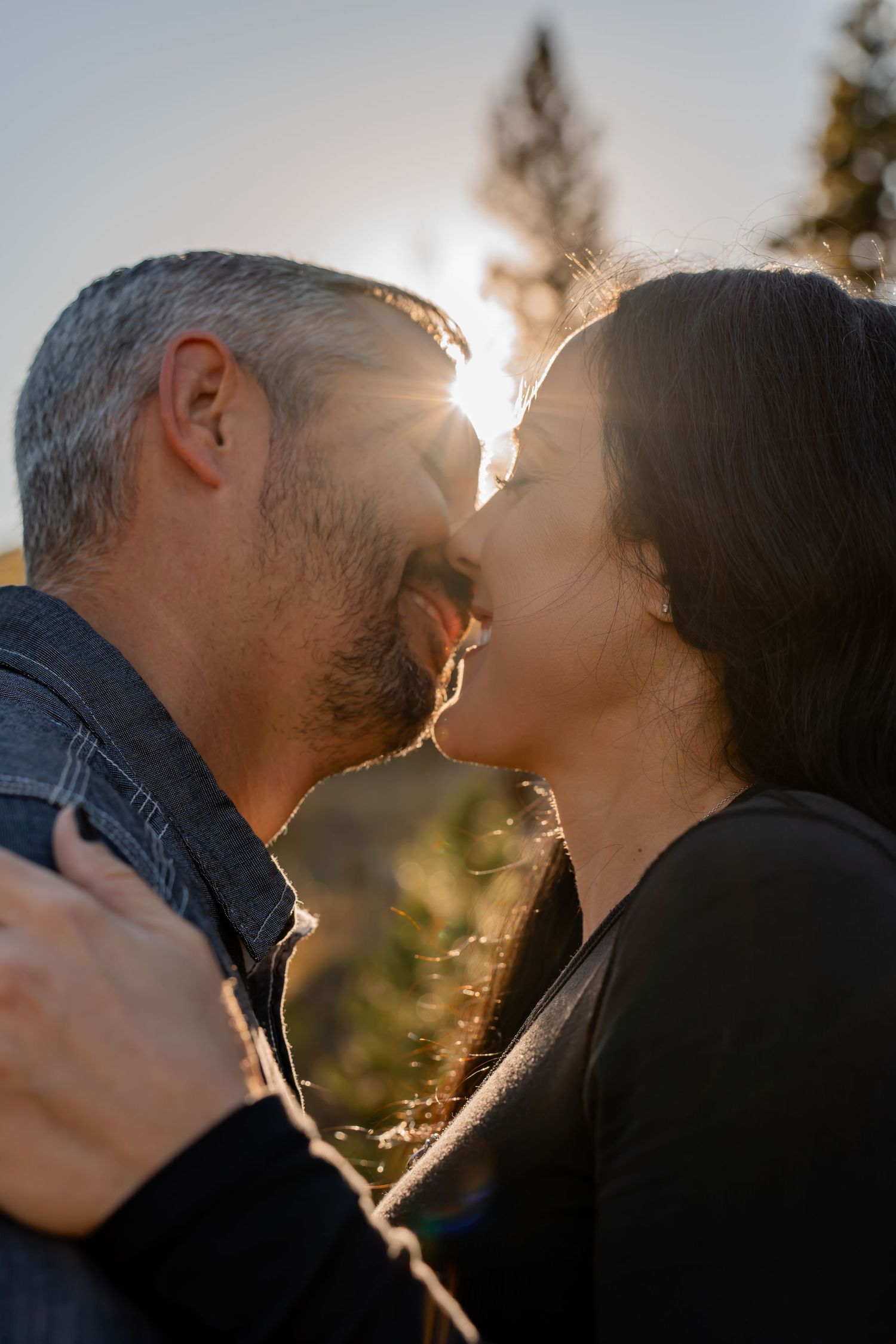 A romantic couple shares an intimate moment at sunset with sunlight creating a warm backlit glow through the trees.