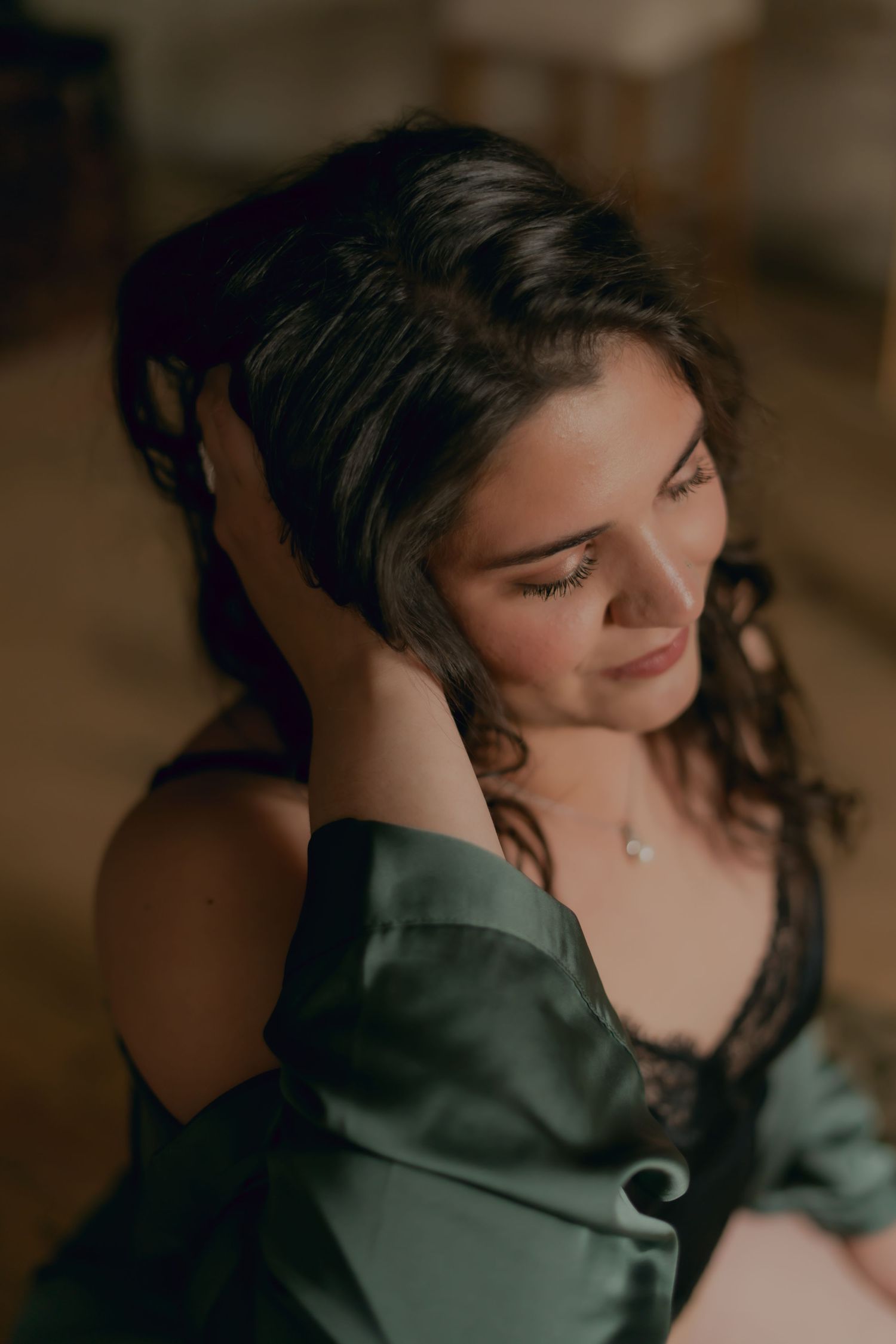 Woman in olive green satin robe gazes down with a soft smile in moody boudoir portrait.
