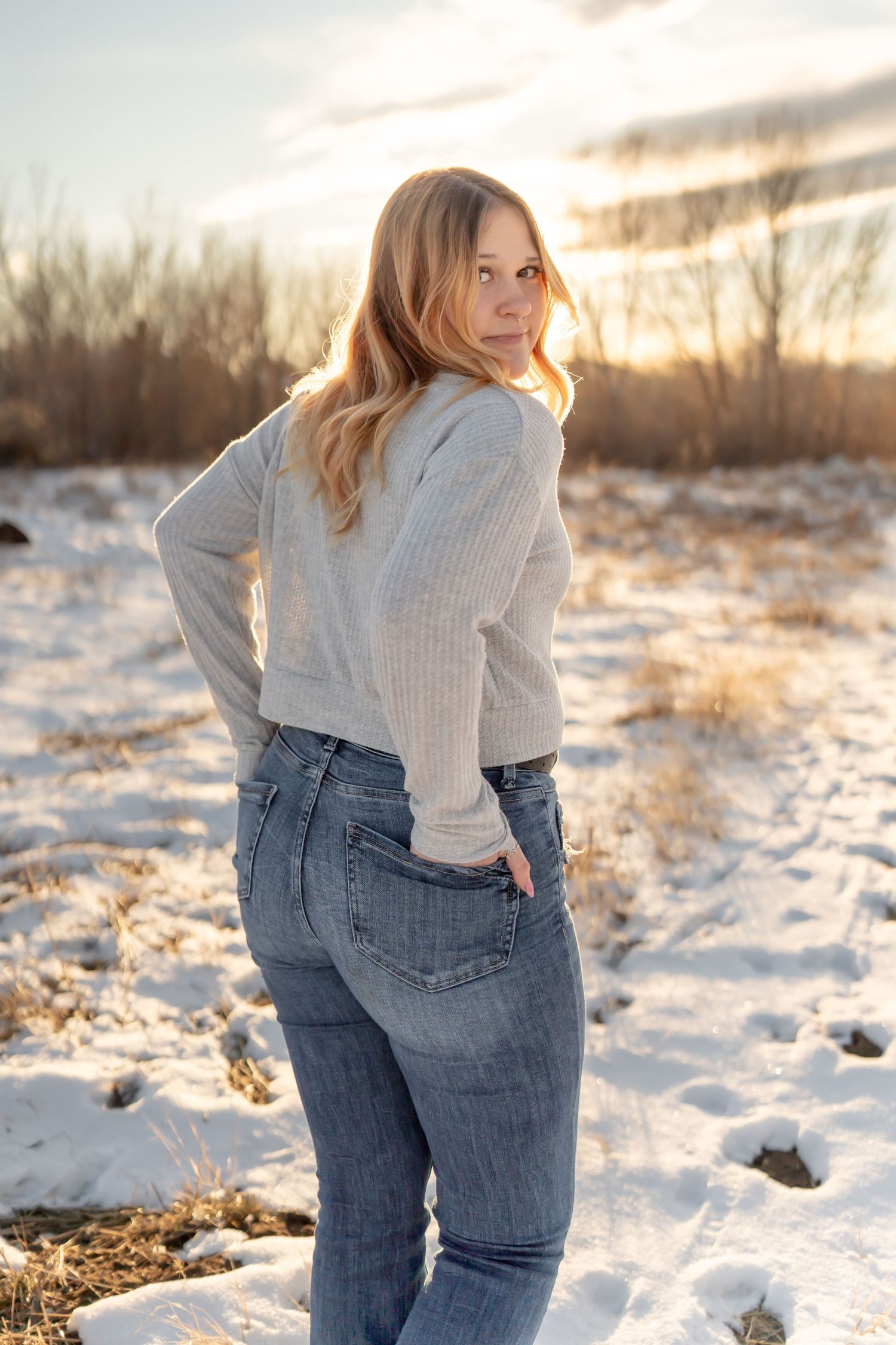 Model wearing light sweater and jeans poses in snowy winter setting during golden hour photoshoot.
