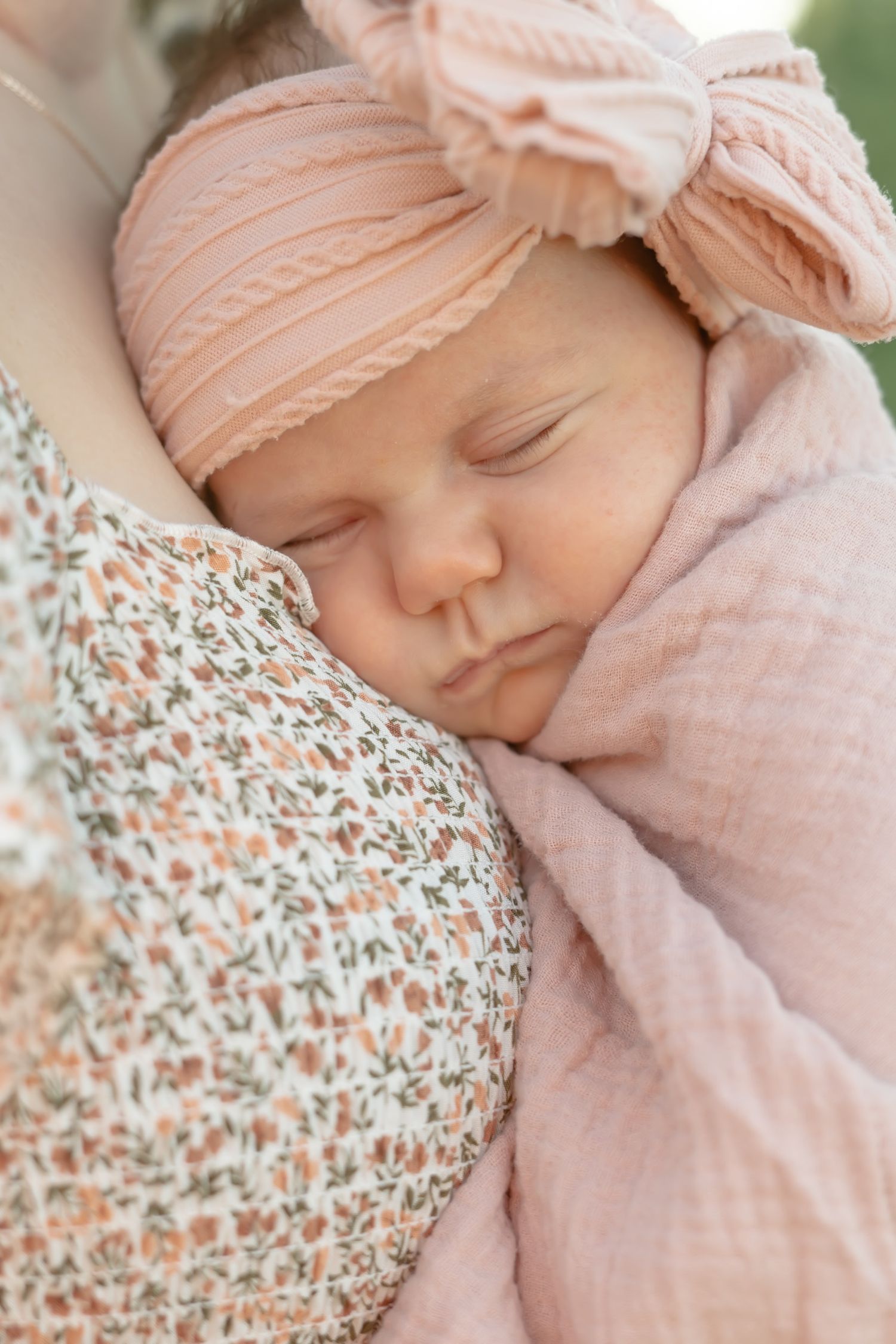 Sleeping baby wearing peach-colored bow headband rests peacefully against floral patterned fabric.
