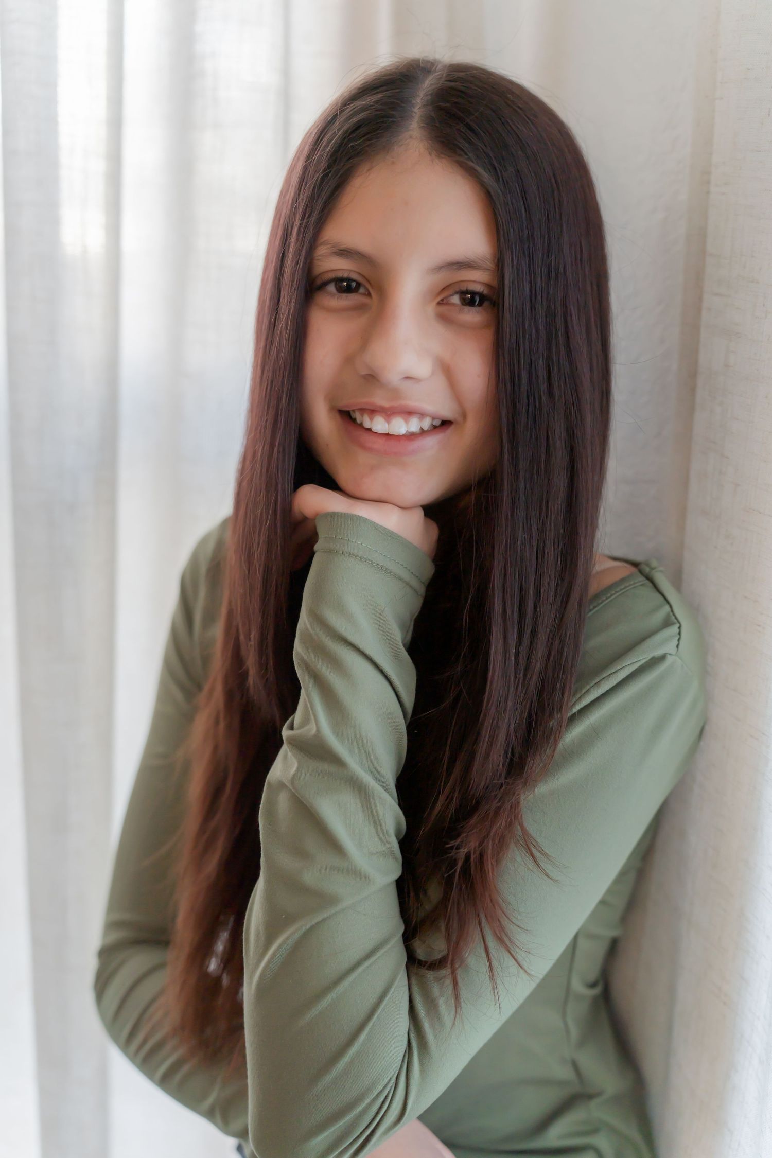 Person with long dark hair wearing a sage green sweater smiles warmly against a light curtained background.