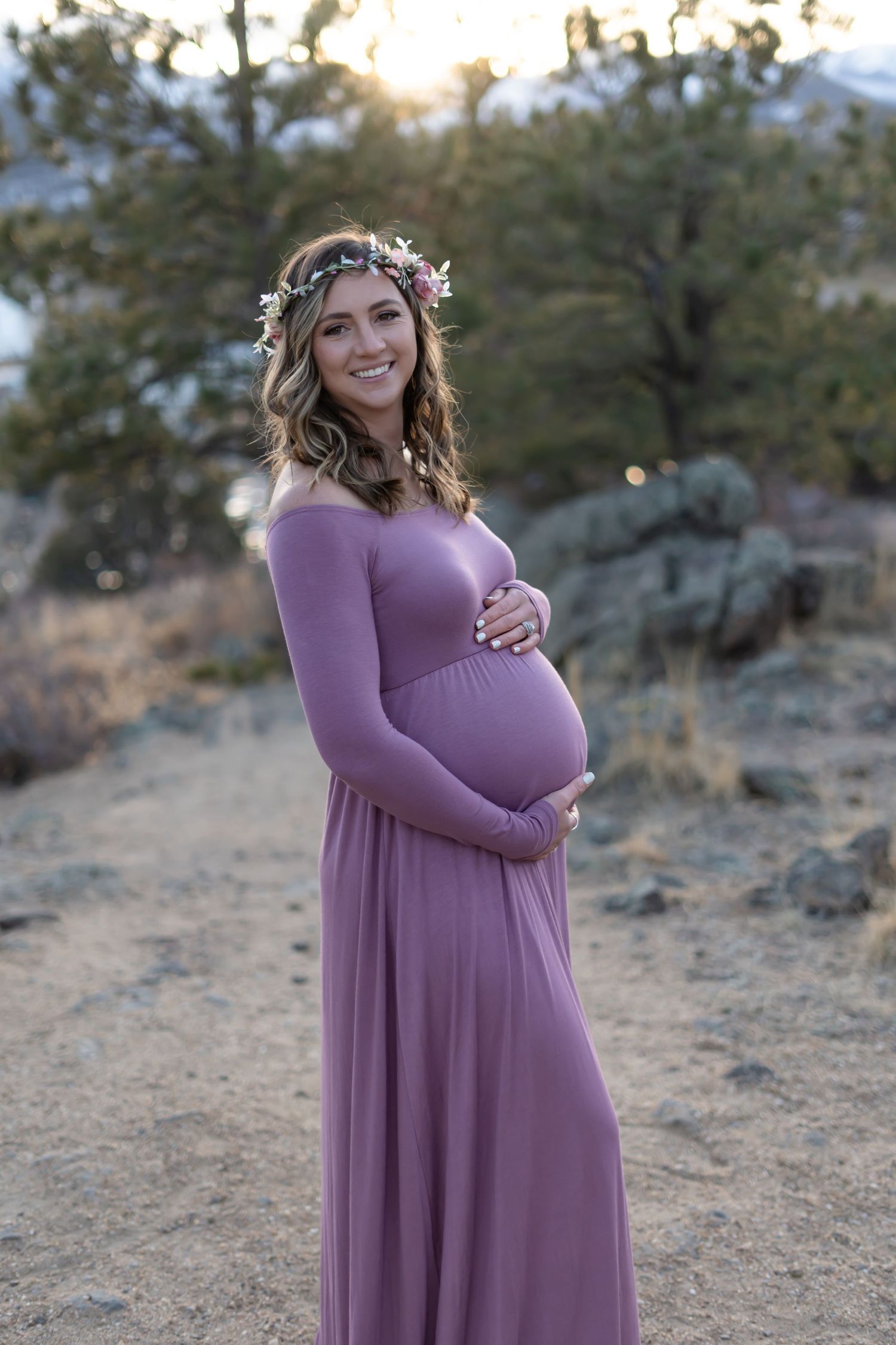 An expectant mother in a flowing purple dress and floral crown poses in a mountain setting at sunset.