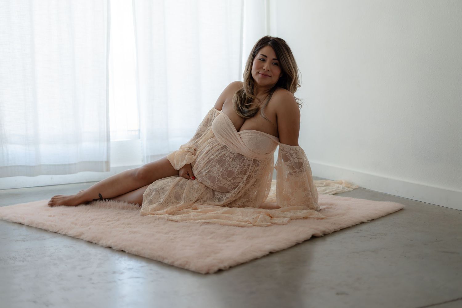 Maternity session with woman in blush dress reclining on floor near window.