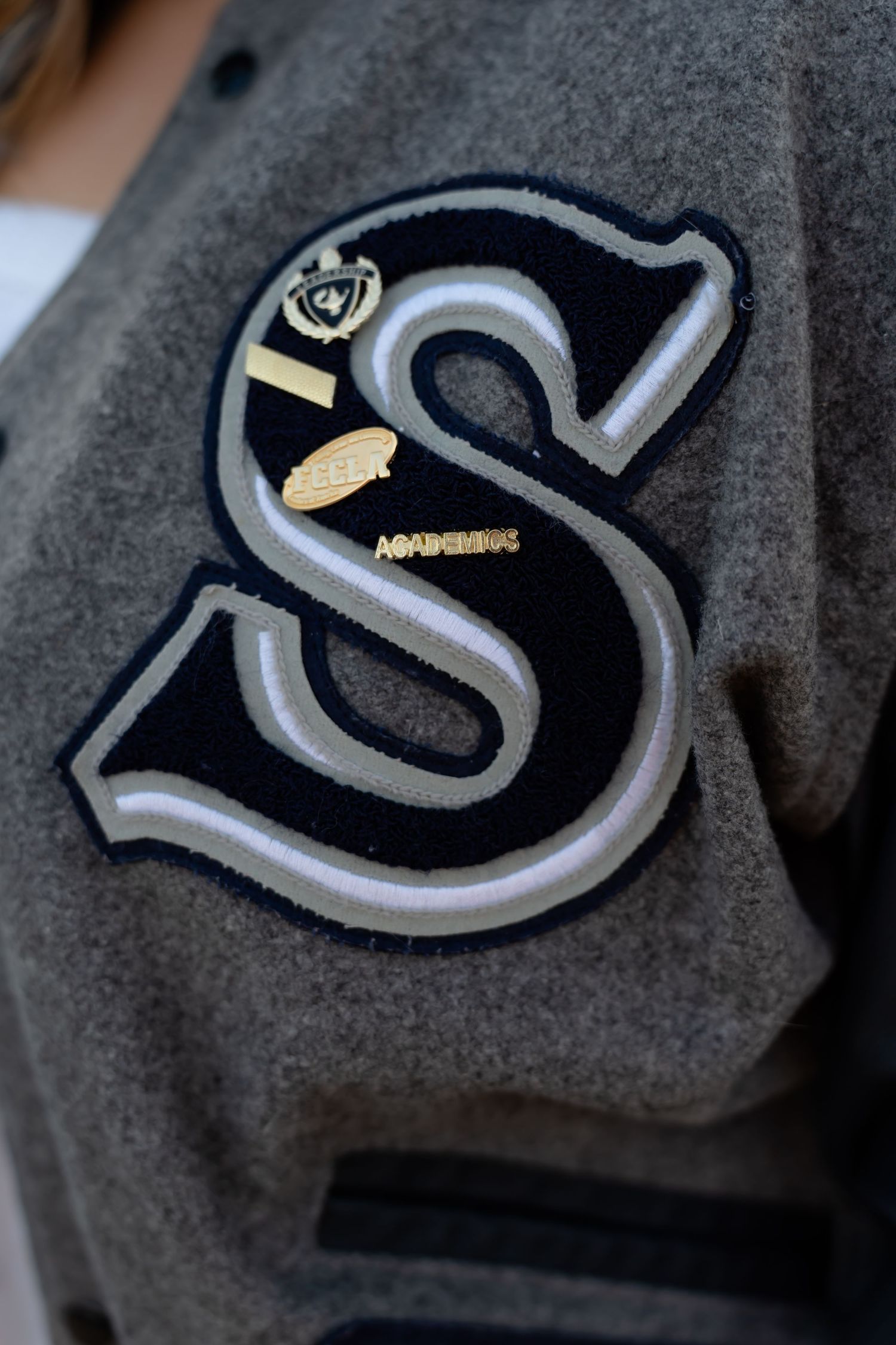 Detailed view of a gray varsity jacket patch with silver and black embroidered lettering.