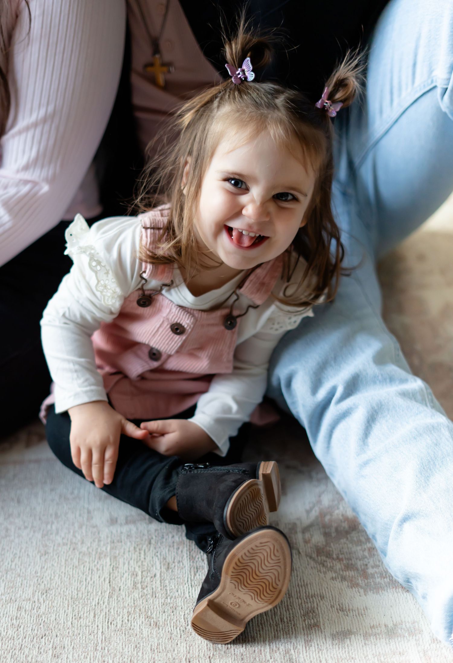 Joyful small child wearing pink overalls and a white shirt sits on carpet with pigtails and a big smile.