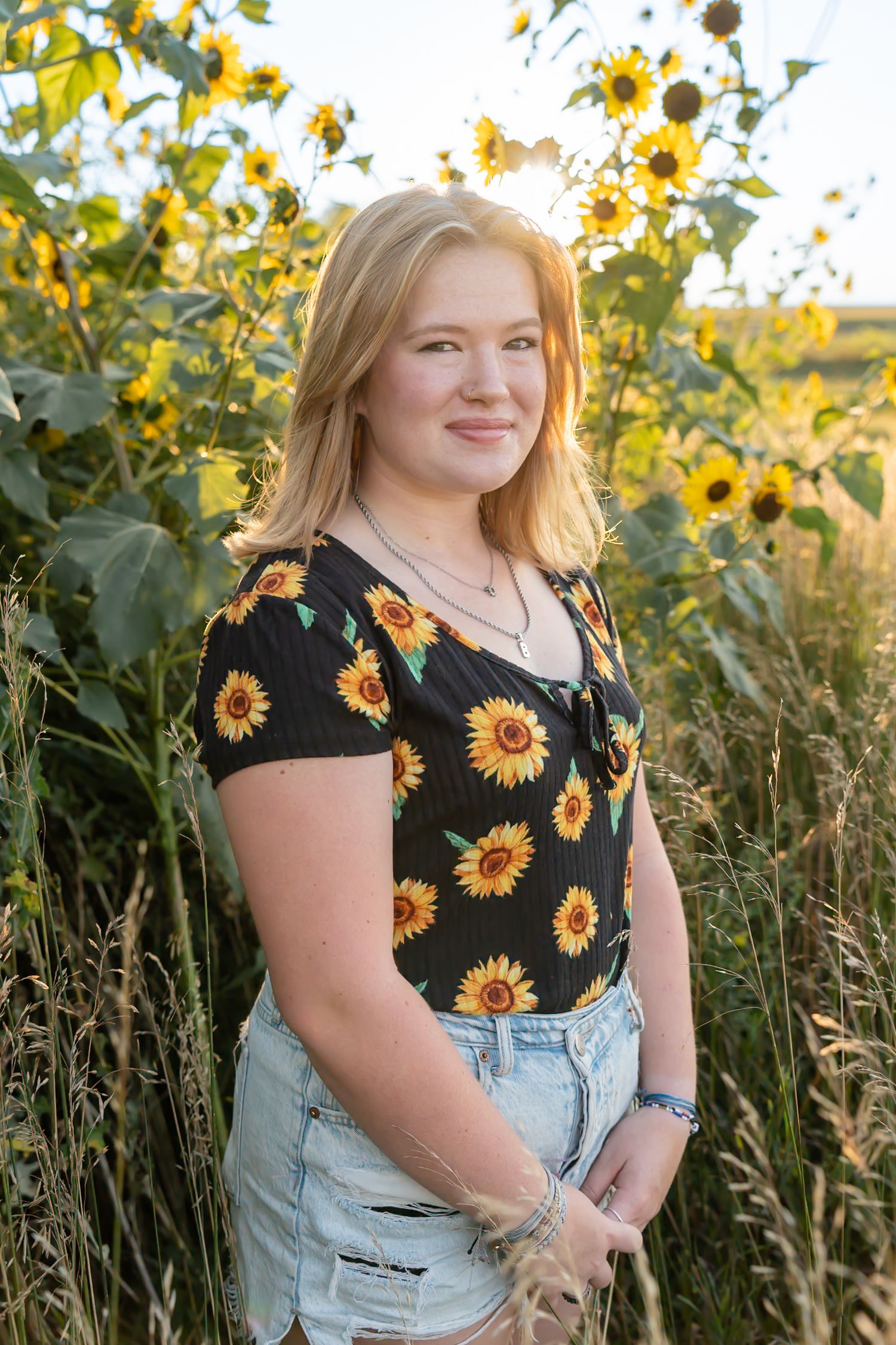 A person wearing a sunflower print top and denim overalls poses in a field of tall sunflowers at sunset.