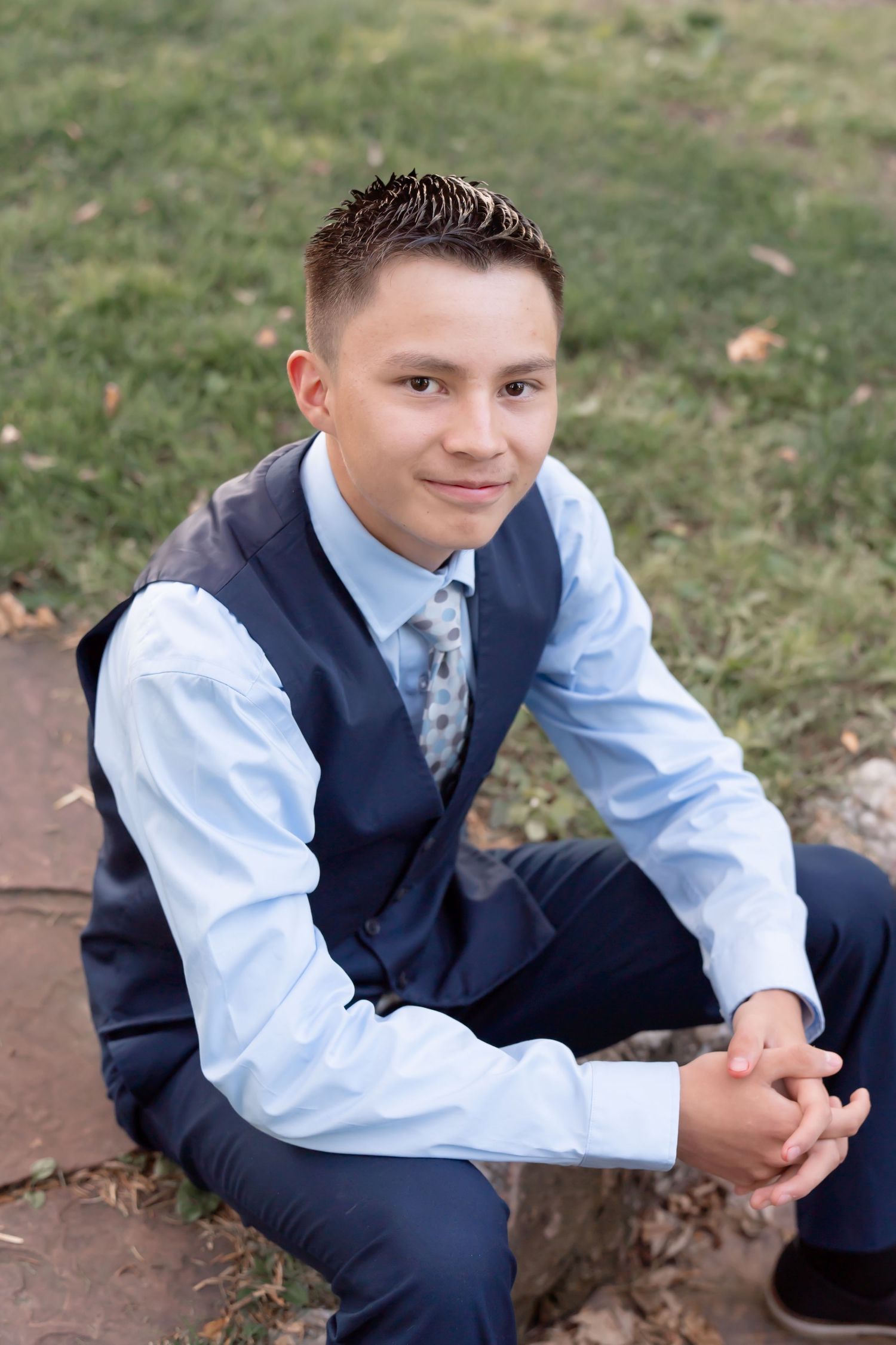 Student in formal navy vest and light blue dress shirt sits casually outdoors.