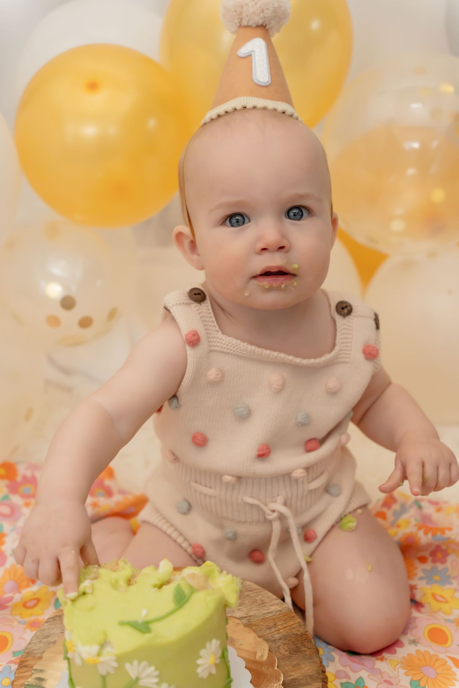 A baby in a cream polka dot outfit celebrates their first birthday with yellow balloons and a green cake.