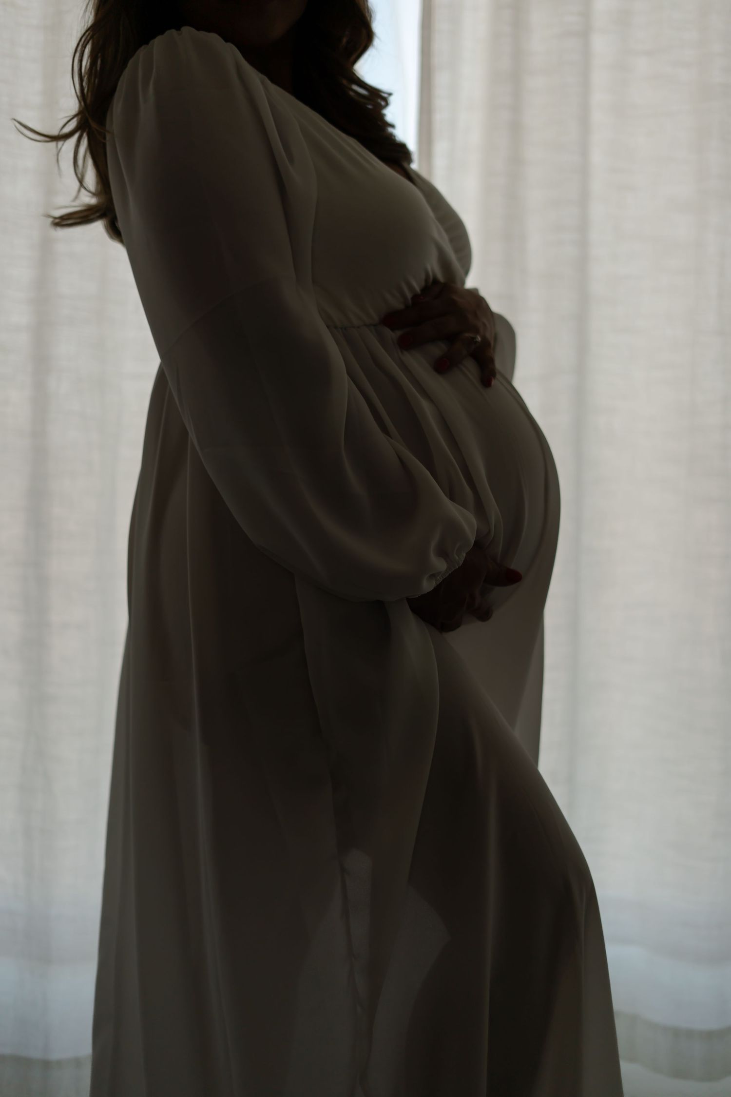 Silhouette of pregnant woman in flowing dress against sheer curtain background.