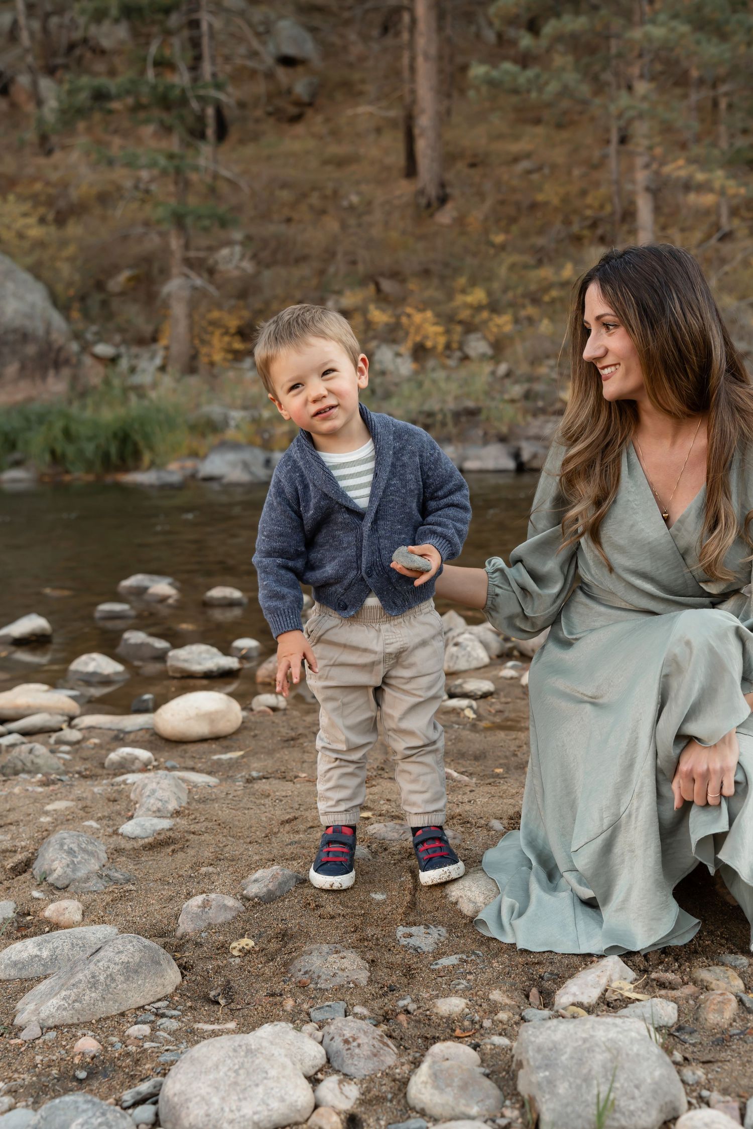 A parent and child sit together on rocks by a lake shore during an outdoor adventure.