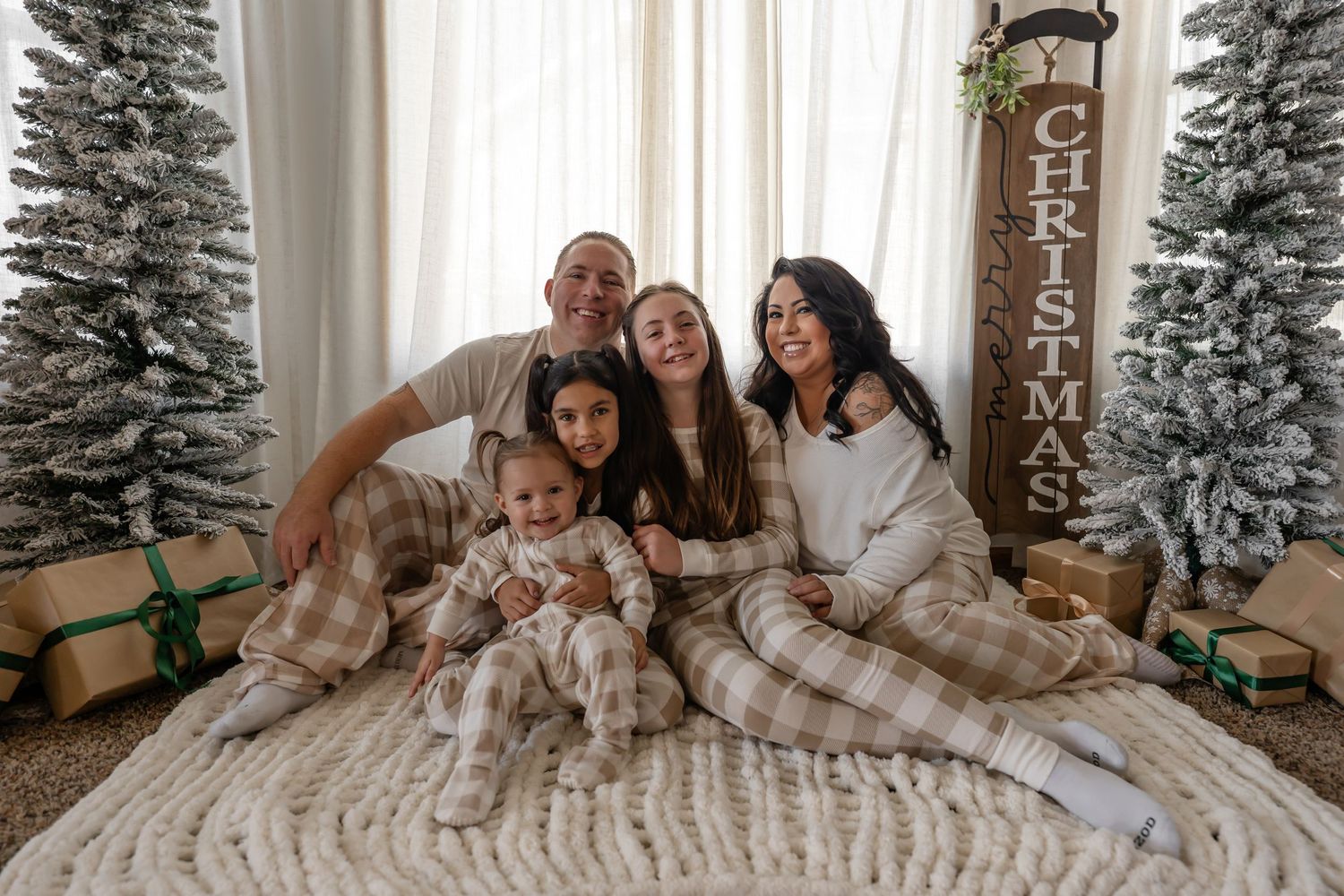 A family wearing matching plaid pajamas sits together on a blanket between snowy Christmas trees and wrapped presents.