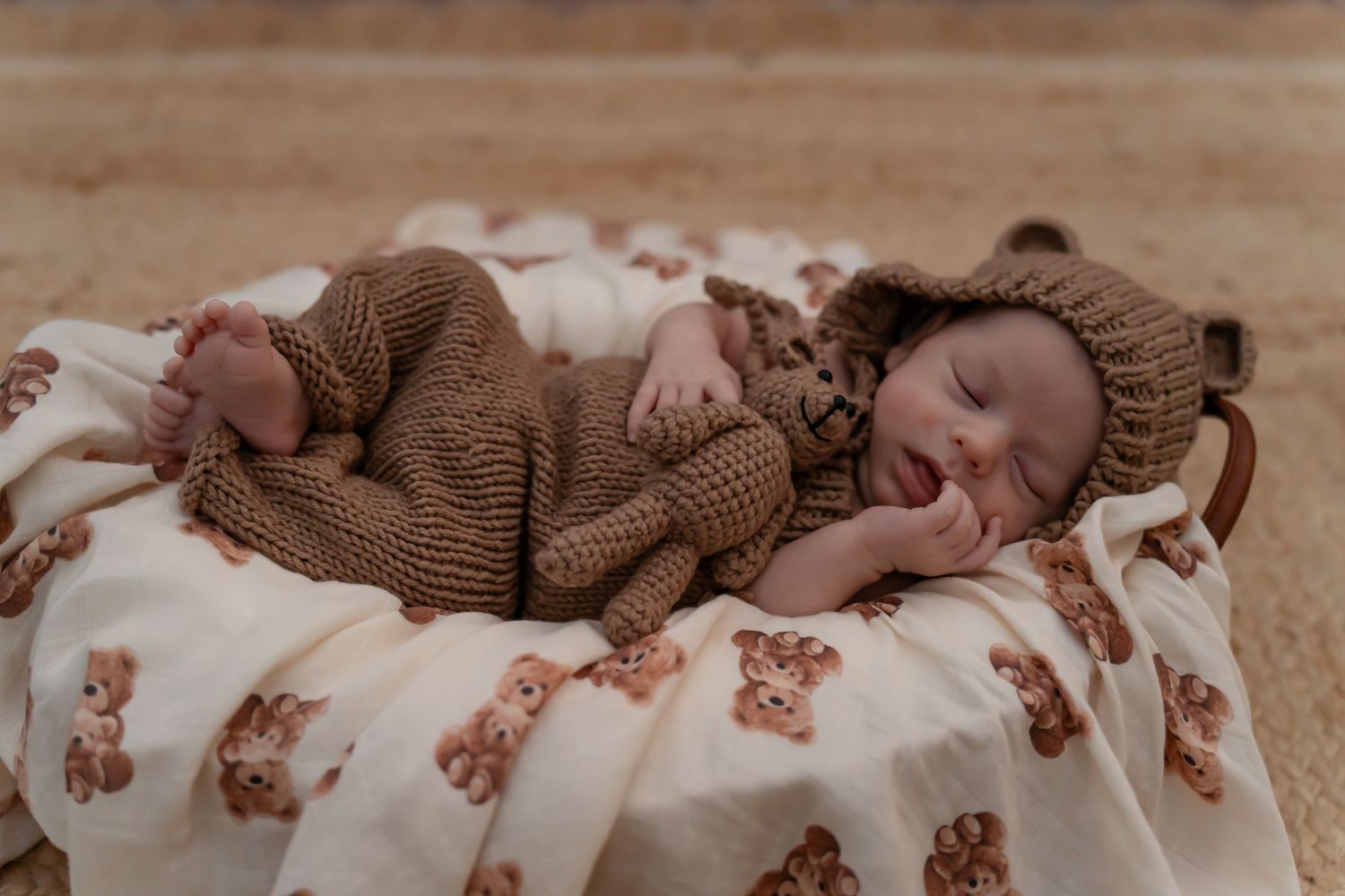 A sleeping baby wearing a brown knit outfit lies peacefully on white bedding with floral patterns.