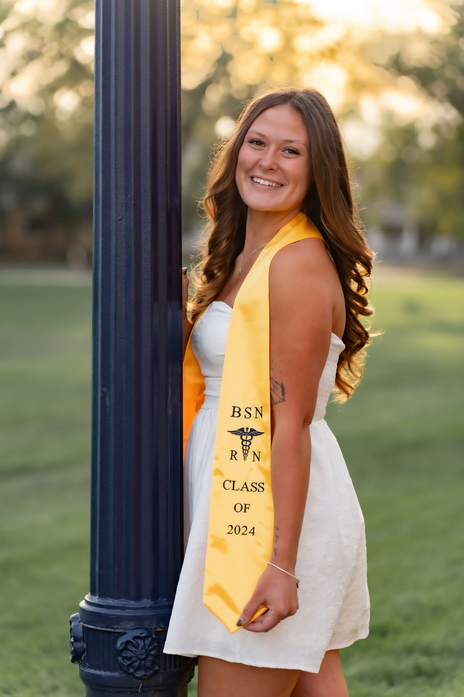 BSN graduate poses in white dress and gold sash with class of 2024 regalia outdoors.